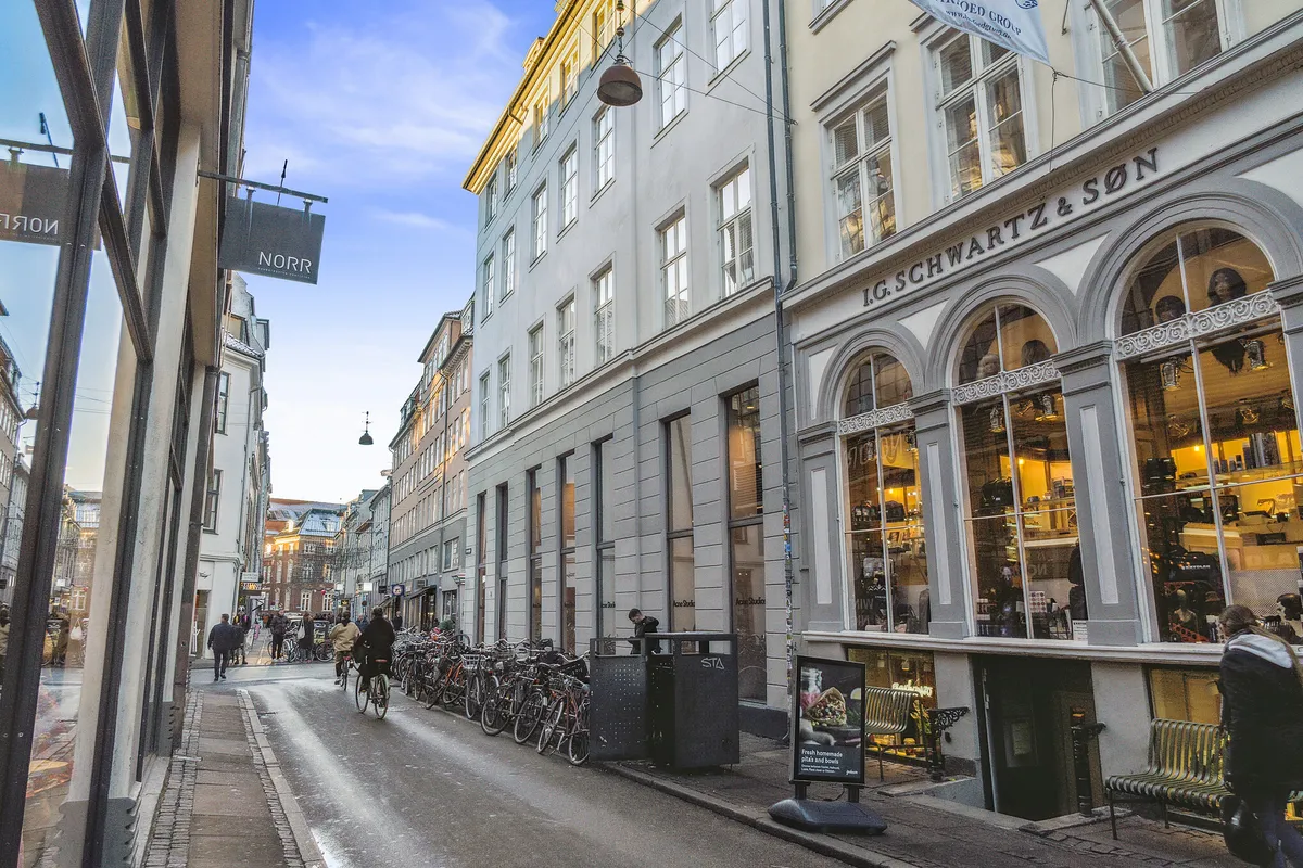 Mennesker går og cykler ad en gågade. Butikker med store ruder. En række cykler står parkeret. Lysskiltene "NORR" og "G. SCHWARTZ & SON"