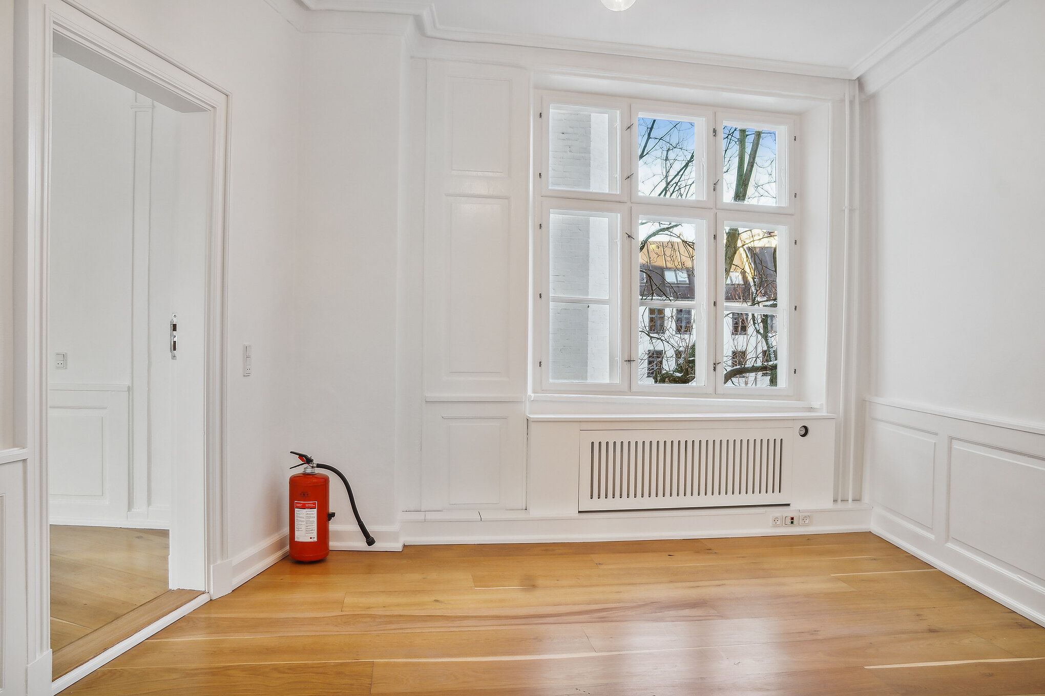 Kontorrum med hvidt panel og gulv, hvor et brandlægskast står mod væggen. Der er et glasvindue med udsigt til bygning og træer udenfor.