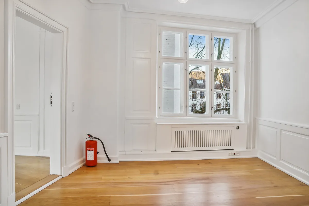 Kontorrum med hvidt panel og gulv, hvor et brandlægskast står mod væggen. Der er et glasvindue med udsigt til bygning og træer udenfor.