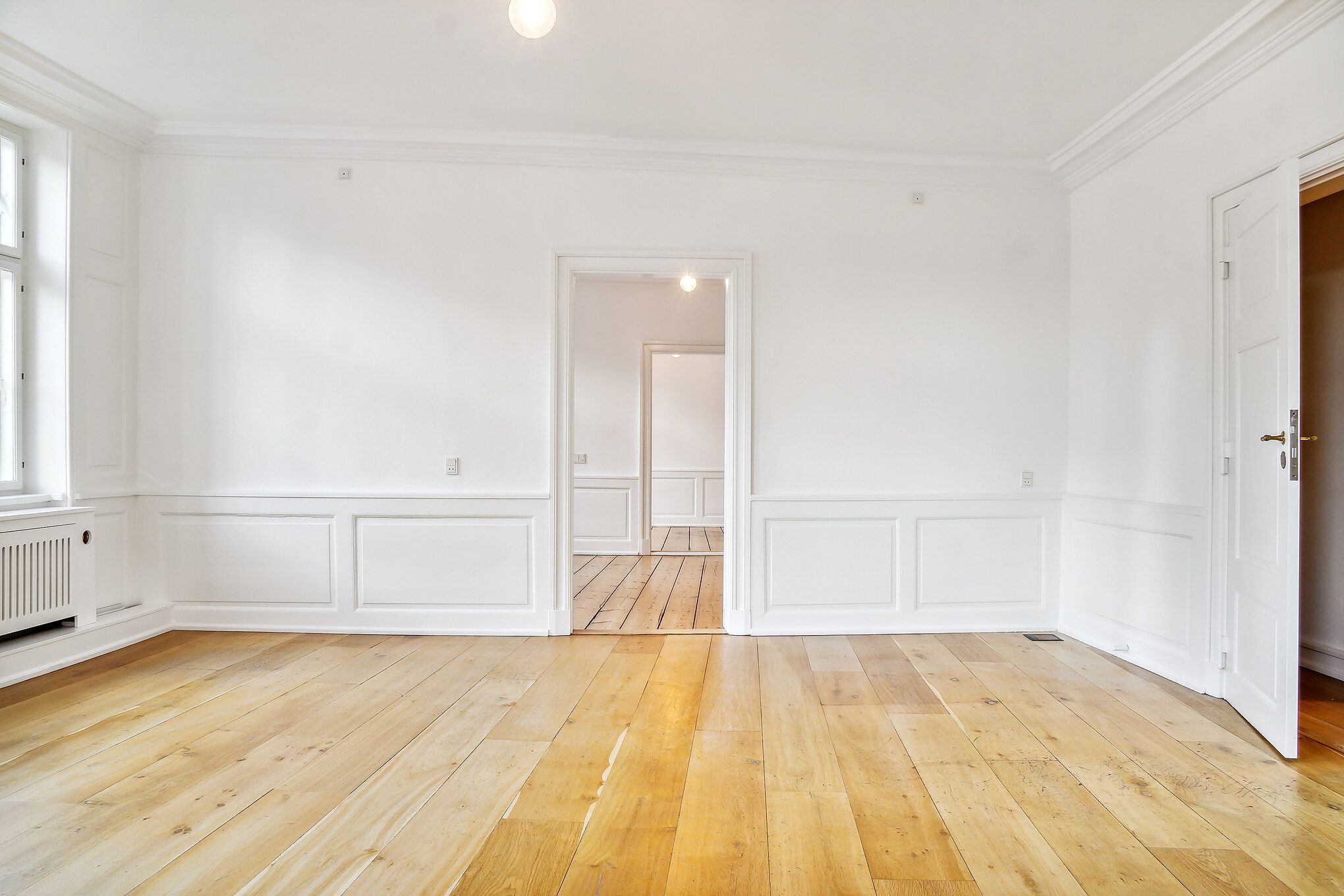 En stor og tom kontorrum med hvidt trægulv og hvide vægge, og lysekroner i loftet. Et kammer og to døre er åbne, og der er et radiator udenfor vinduet