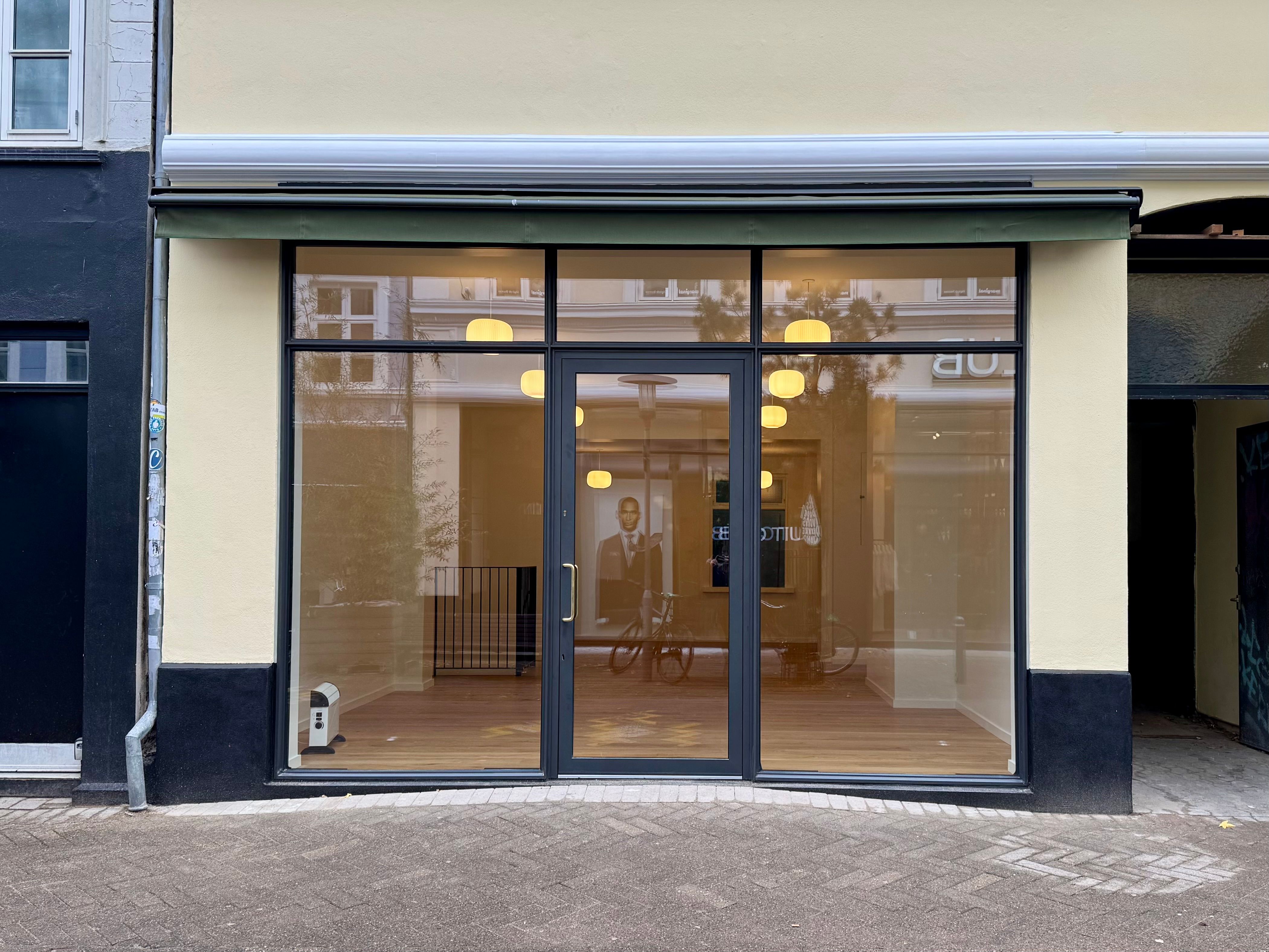 Butiksfront med to store glasdøre og flere vinduer, udsmykket med lysekroner og refleksioner af en mand i ansigt og cykel.