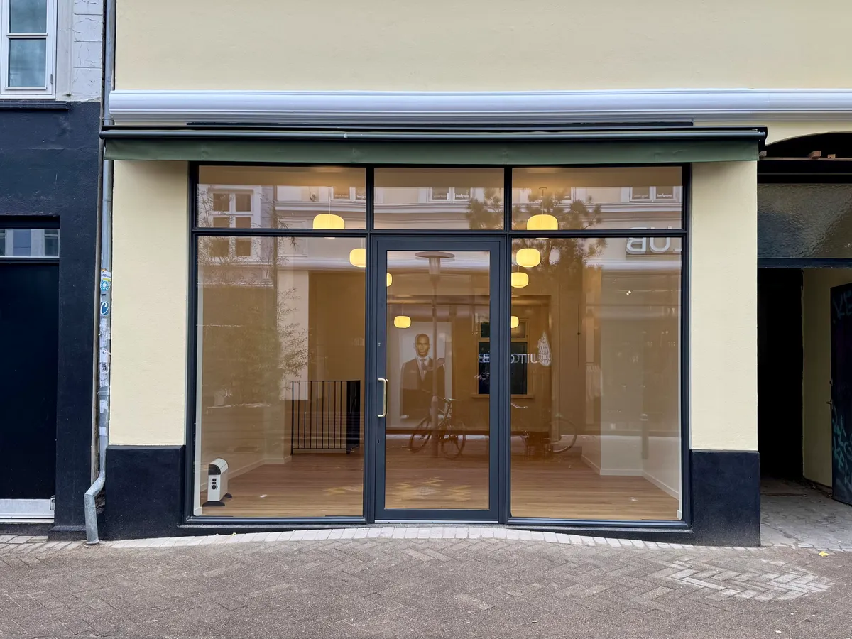 Butiksfront med to store glasdøre og flere vinduer, udsmykket med lysekroner og refleksioner af en mand i ansigt og cykel.