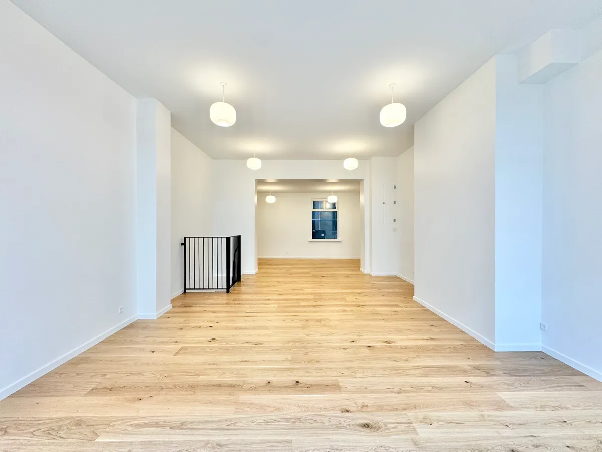 En tom rummelighed med hvide vægge og lyse loftslamper, massivt trægulv, og en slank metalgeværd, der fører til et vinduesområde.