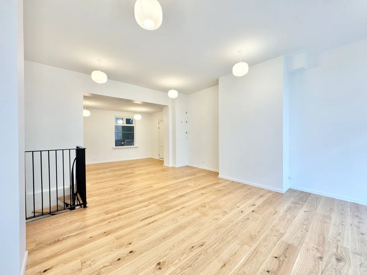 En tom og møbleret butik med hvide vægge og lyse trægulve. Loftet er hvidt med fire hængende lamper og der er en sort stige mod højre.