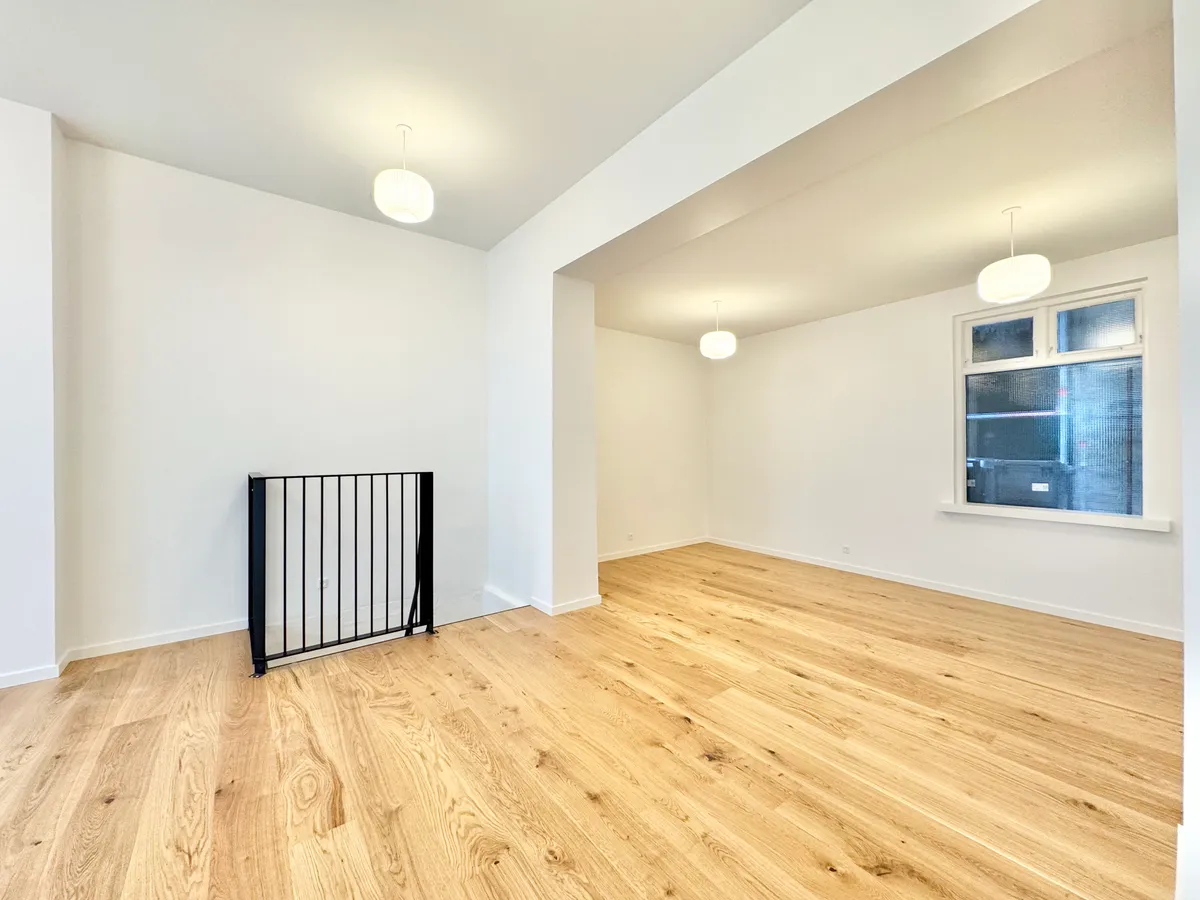 En stor, tom butikslokal med lyse trægulve, hvide vægge og hængelampe på loftet. Vinduet og døren er lukket. På gulvet står en slank, sort og sølvfarvet stang.