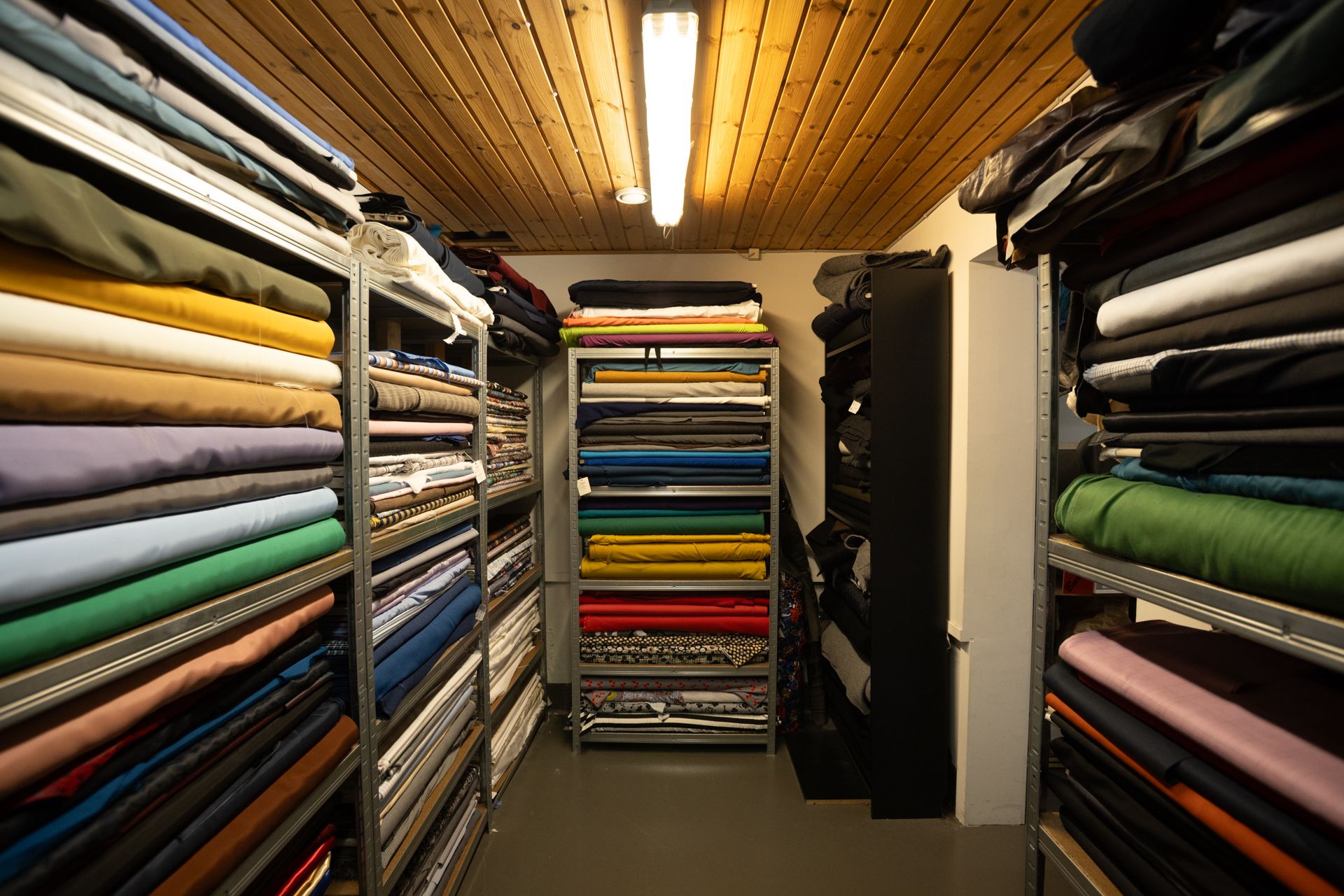 Sortiment af forskellige stoftyper, blå og grønne, på høje hylder i en mørk og varm indretning med lyse loftslampe