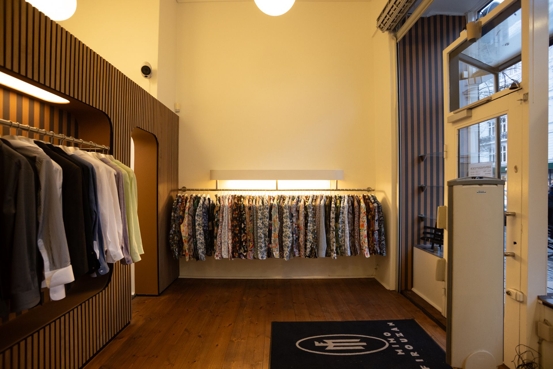 Indkøbsmuligheder på Frederiksborggade 37 i København. Moderne indretning med hylder og tøjstative. Sort gulvtæppe med mærkelogo. Glasset dør og rekvisitter til salg.