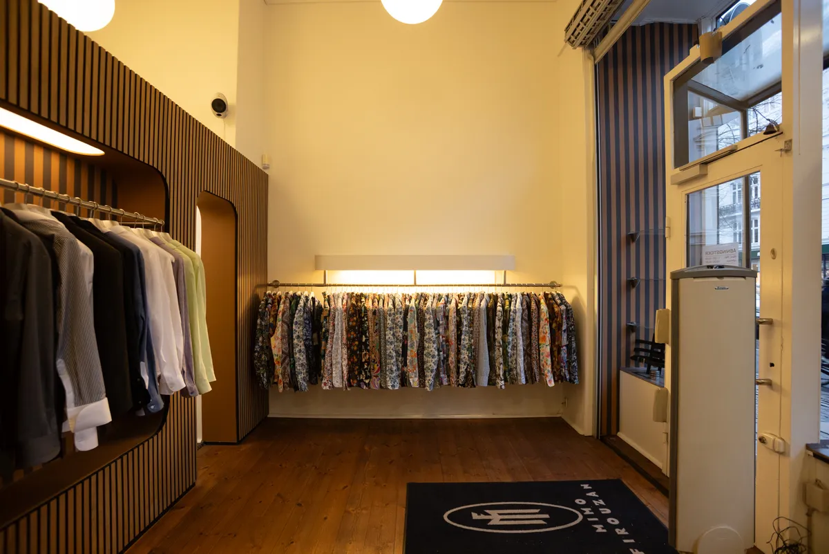 Indkøbsmuligheder på Frederiksborggade 37 i København. Moderne indretning med hylder og tøjstative. Sort gulvtæppe med mærkelogo. Glasset dør og rekvisitter til salg.