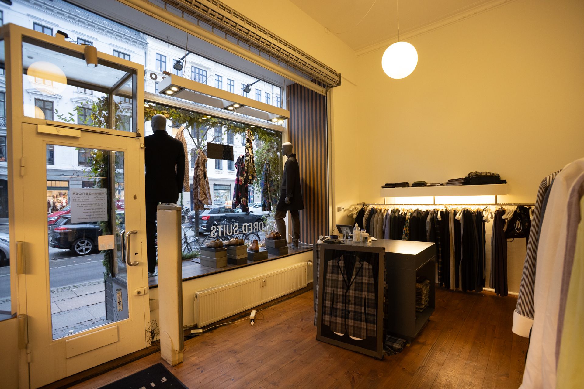 Foto af en lukket butik med trægulv og lyse pupper, hvor der er hængende tøj og en stol med en jakke