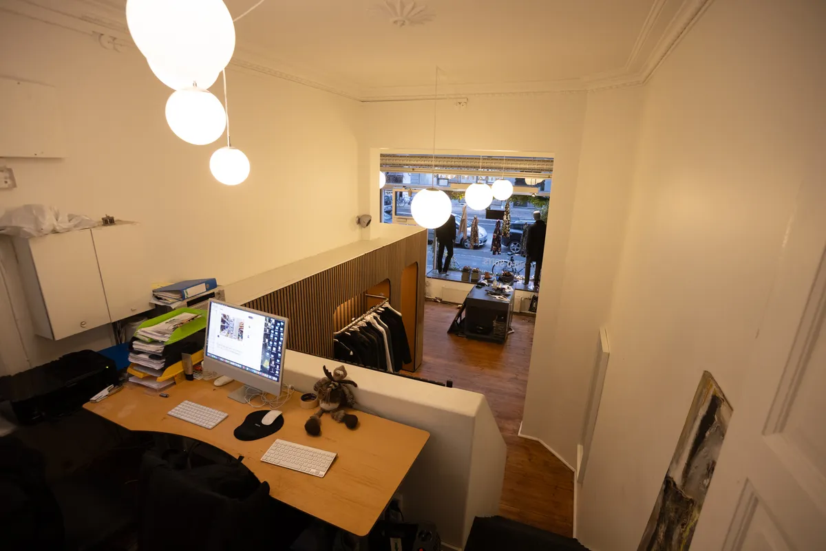 Et arbejdsrum med en skærm og en computer, et tæppe og en møbelsektion, hvor der hænger tøj, og en åben dør med udsigt til gaden.