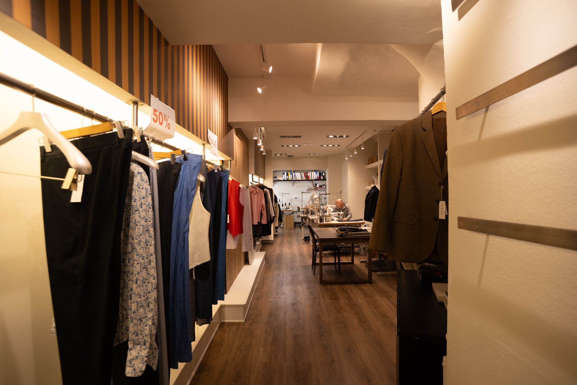 Tøjreol med 50% rabat, trægulv og lysekroner i et stilfuldt boutique med træhylder og kunder og medarbejdere i baggrunden