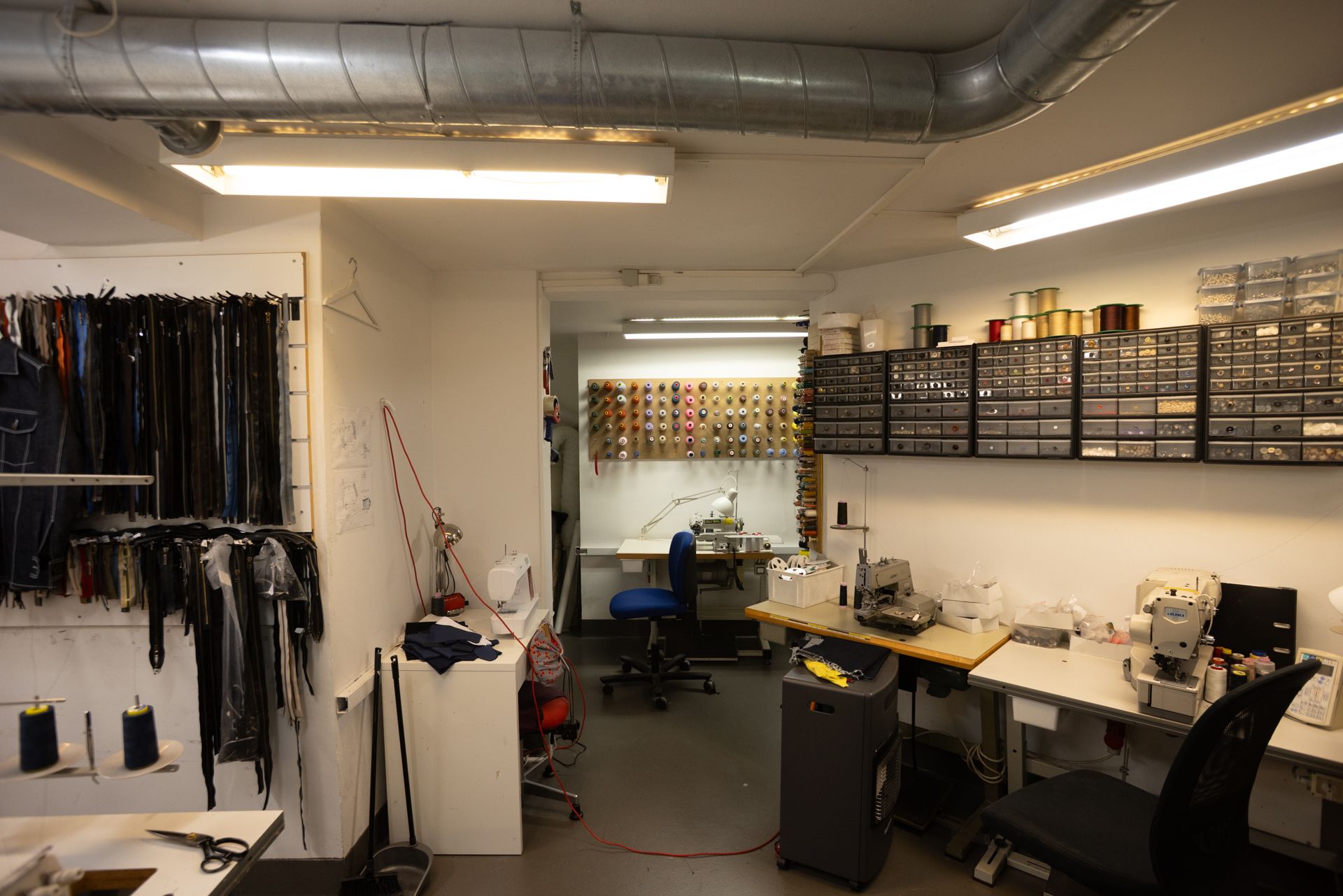 Lejlighed med syværksted og tilbehør på Frederiksborggade 37. Forskellige maskiner og stoffer. Sorte stole og lyse loftslampe. Sorte linjer og søm i væggen.