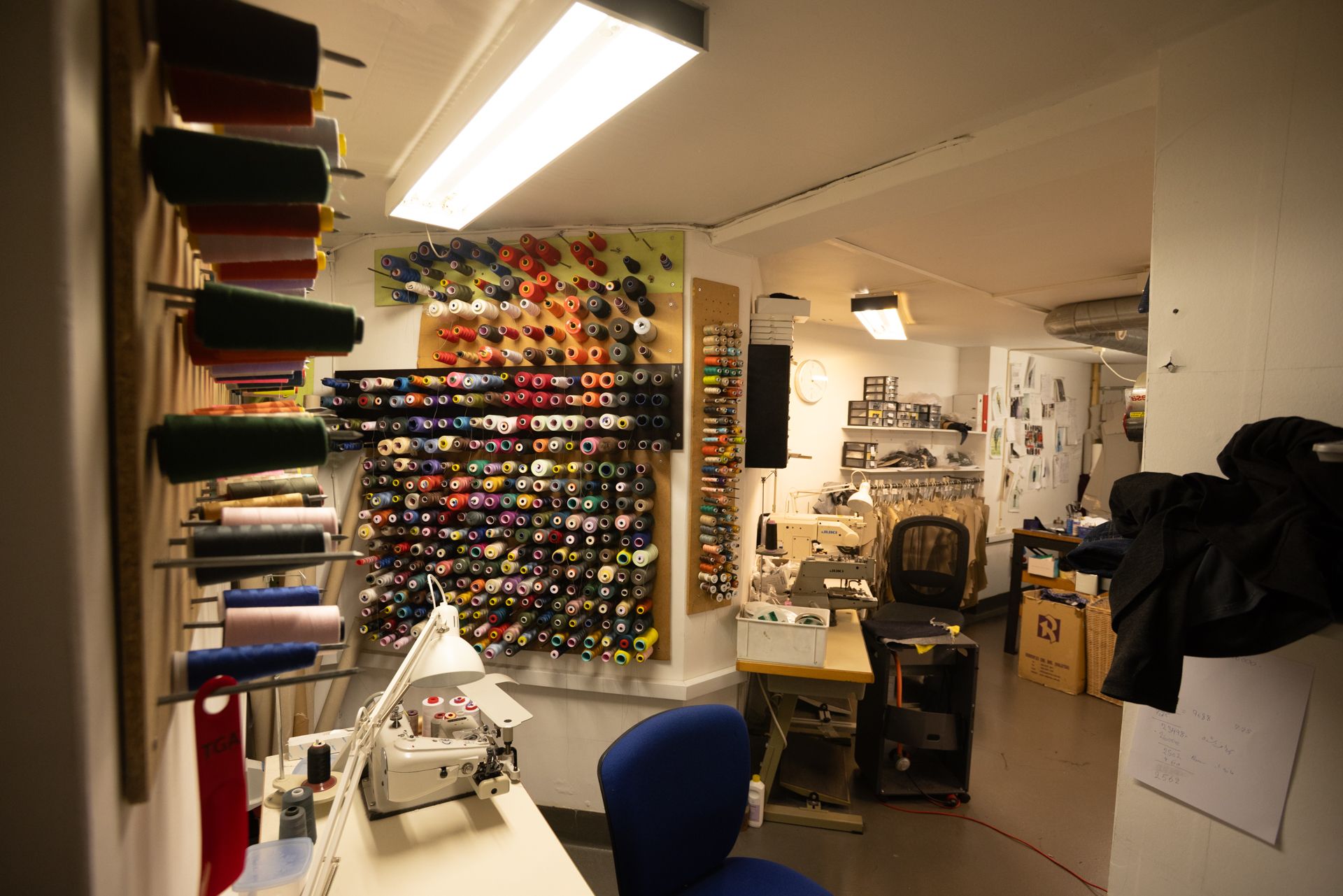 En butik med sømmemaskine og skærestol, samt flere farverige trådspoler, og en række sømmemaskiner. Der er flere sømmemaskiner og en række kasser på gulvet