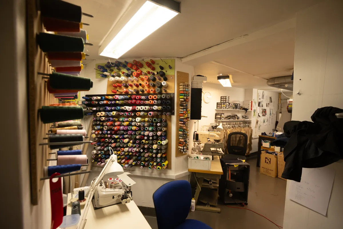 En butik med sømmemaskine og skærestol, samt flere farverige trådspoler, og en række sømmemaskiner. Der er flere sømmemaskiner og en række kasser på gulvet