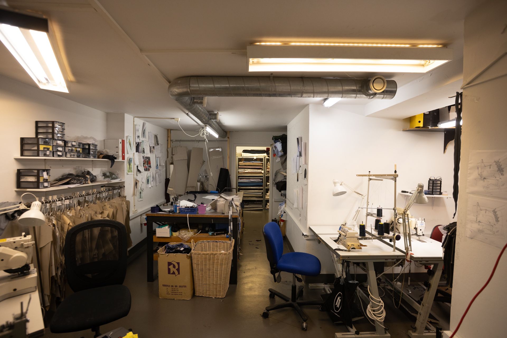 Billede af et værksted med et stort arbejdebord, søjler, hængende tøj og en stol samt flere søjler og hylder med tøj og andre ting