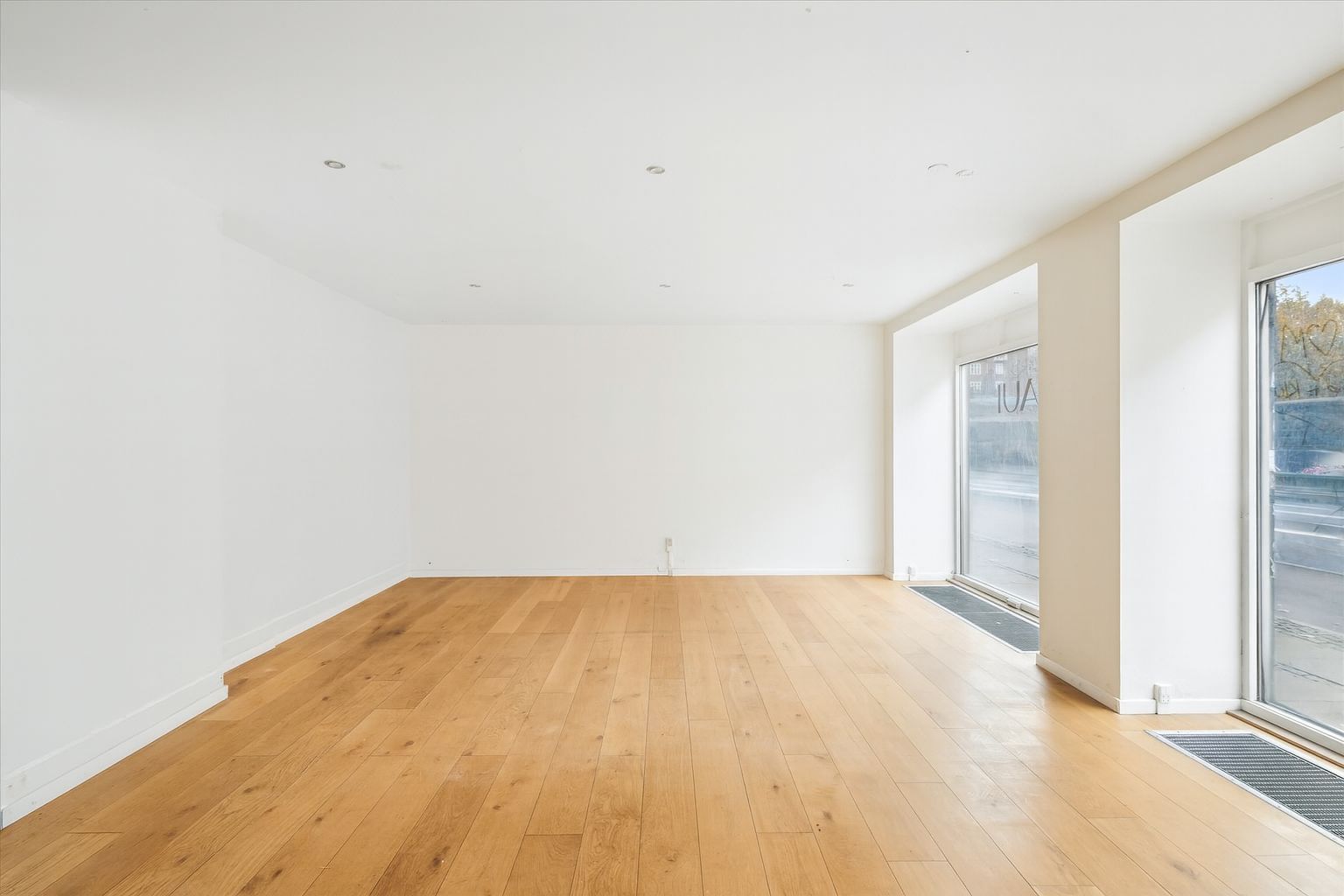 Et stort, lyse og pænt erhvervslokale med trægulve, hvide vægge og store glasruder mod gaden og et hjørne.