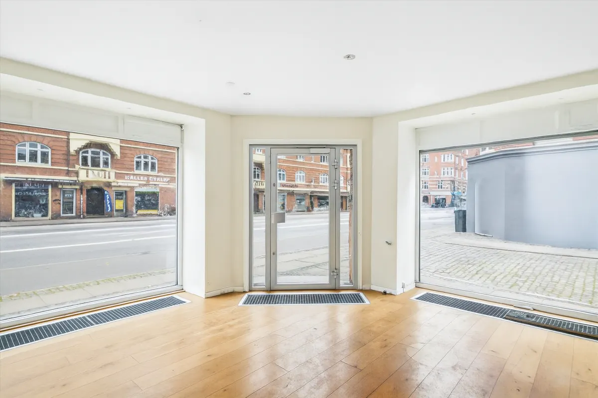 Erhvervslokale med store glasvinduer og hjørnefacade, lyse indvendige vægge og trægulve, og en udsigt ud mod Amagerbrogade og krydset med en smal gade