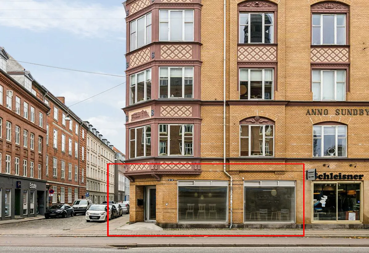 En stor, rødmalet bygning med flere vinduer og et hjørne i Amagerbrogade, København. To butikker med glasvinduer og teksten Schleisner i hjørnet.