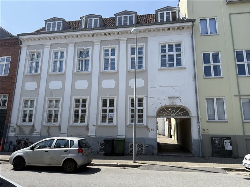 En hvid bygning med vinduer, indgang og nummer 34 i Fredericia. En sølvfarvet bil er parkeret foran bygningen.