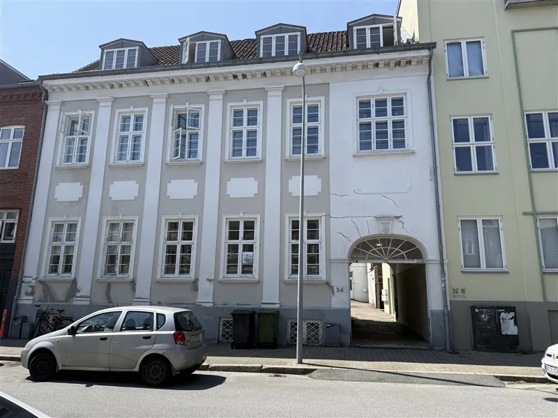 En hvid bygning med vinduer, indgang og nummer 34 i Fredericia. En sølvfarvet bil er parkeret foran bygningen.