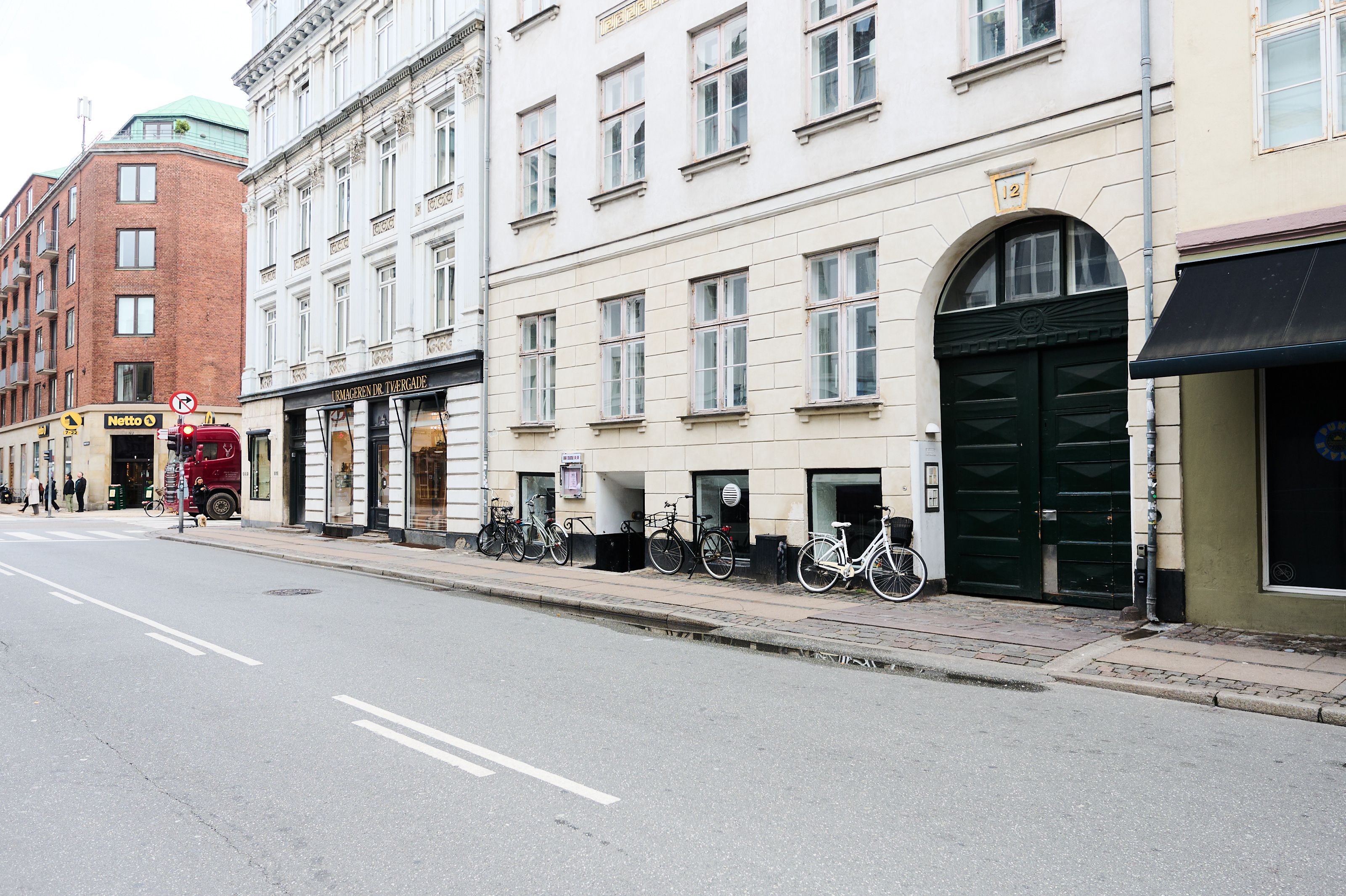 Vej med cykler og biler i udsigt. En bygning med en nummerplade og en butiksdør i København. En rødt lastbil og en person står på gaden.