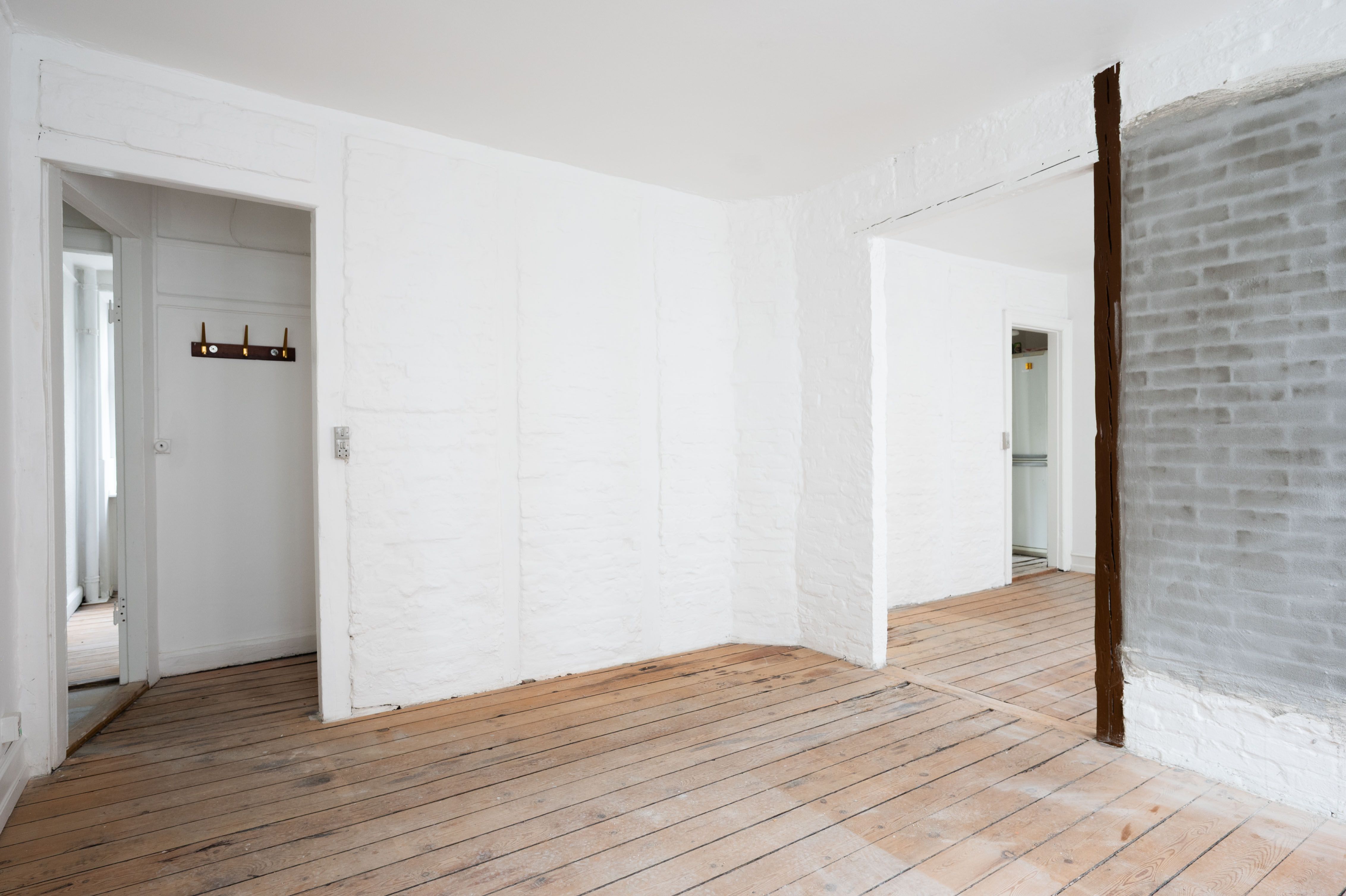 Et tomt rum med hvide vægge og lyse trægulve samt lysepunkter og en åben dør til et lille rum med hvide vægge og en åben dør