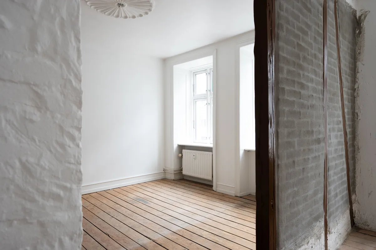 Foto af en tom og renoveret lejlighed med hvide vægge og trægulv, to vinduer med hvide rammer, og en åben dør til en ny opført væg med stenflis