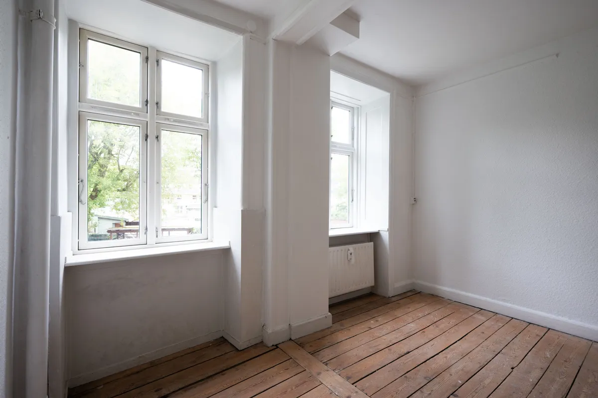 En værelse med hvide vægge og gulvtæppe, to vinduer med træ udvendigt og et radiator mellem vinduerne