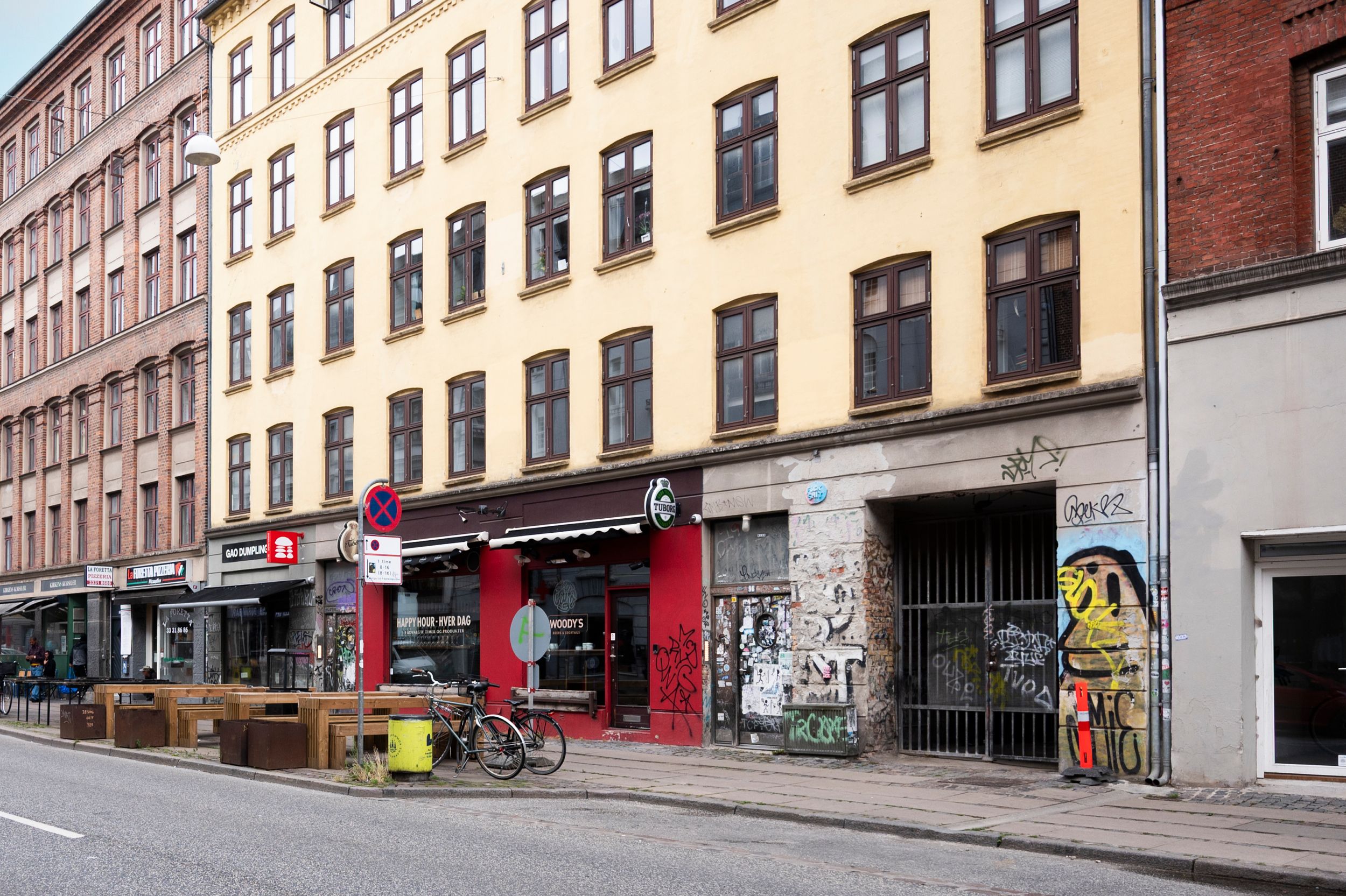 En bygning med flere etager, graffiti og vægmaleri. Cykler står parkeret foran, og der er et gult spildevandsfad på fortovet.