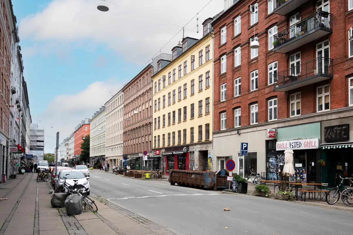 Gade med mange bygninger og forretninger, der er udsmykket med terrasser og udsigt over gaden. Parkerede biler og cykler samt udskrævede skilte og forretningsviser.
