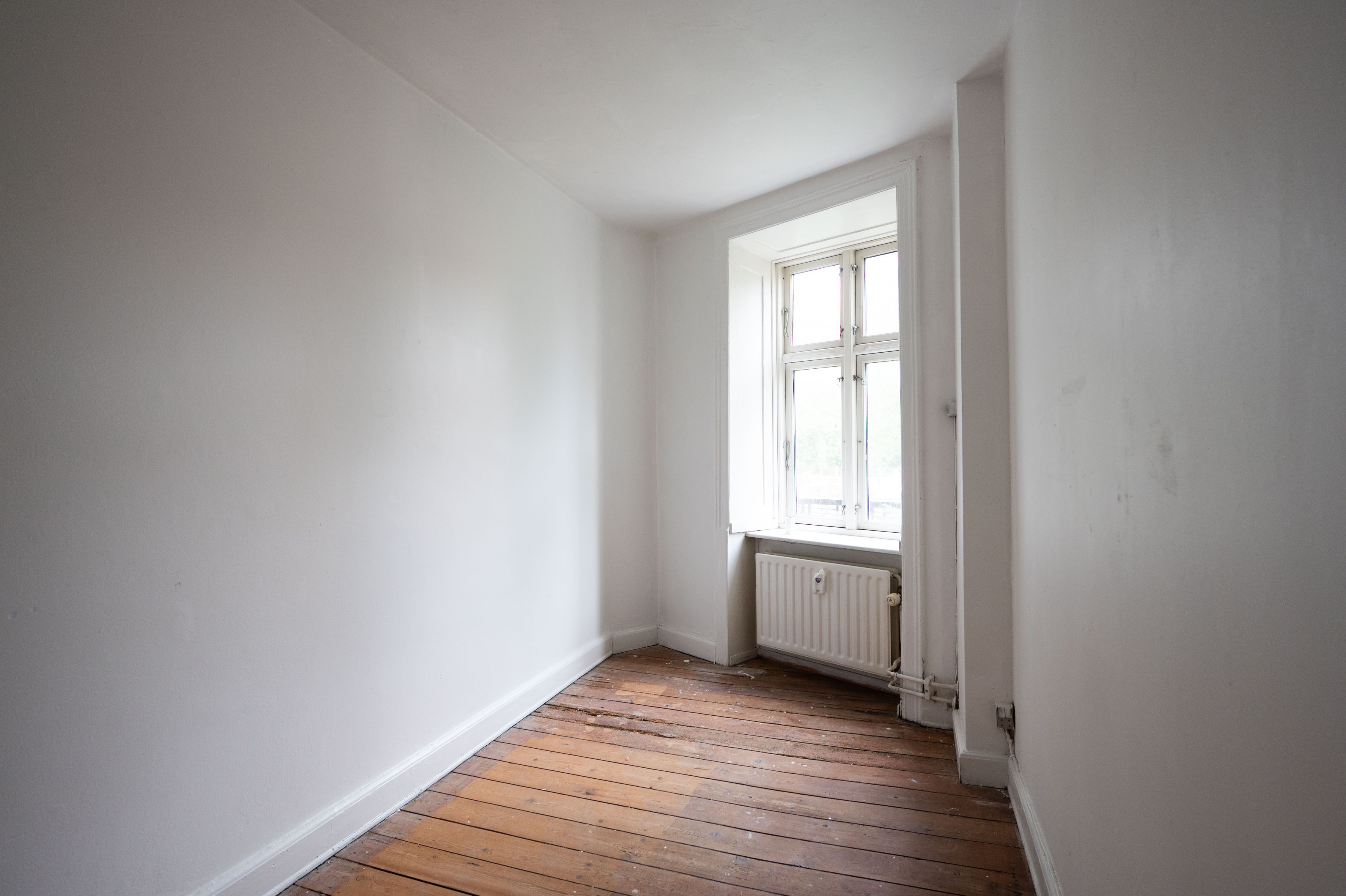 En tom stue med hvide vægge og lyse trægulve, samt en vinduesåbning med radiator.