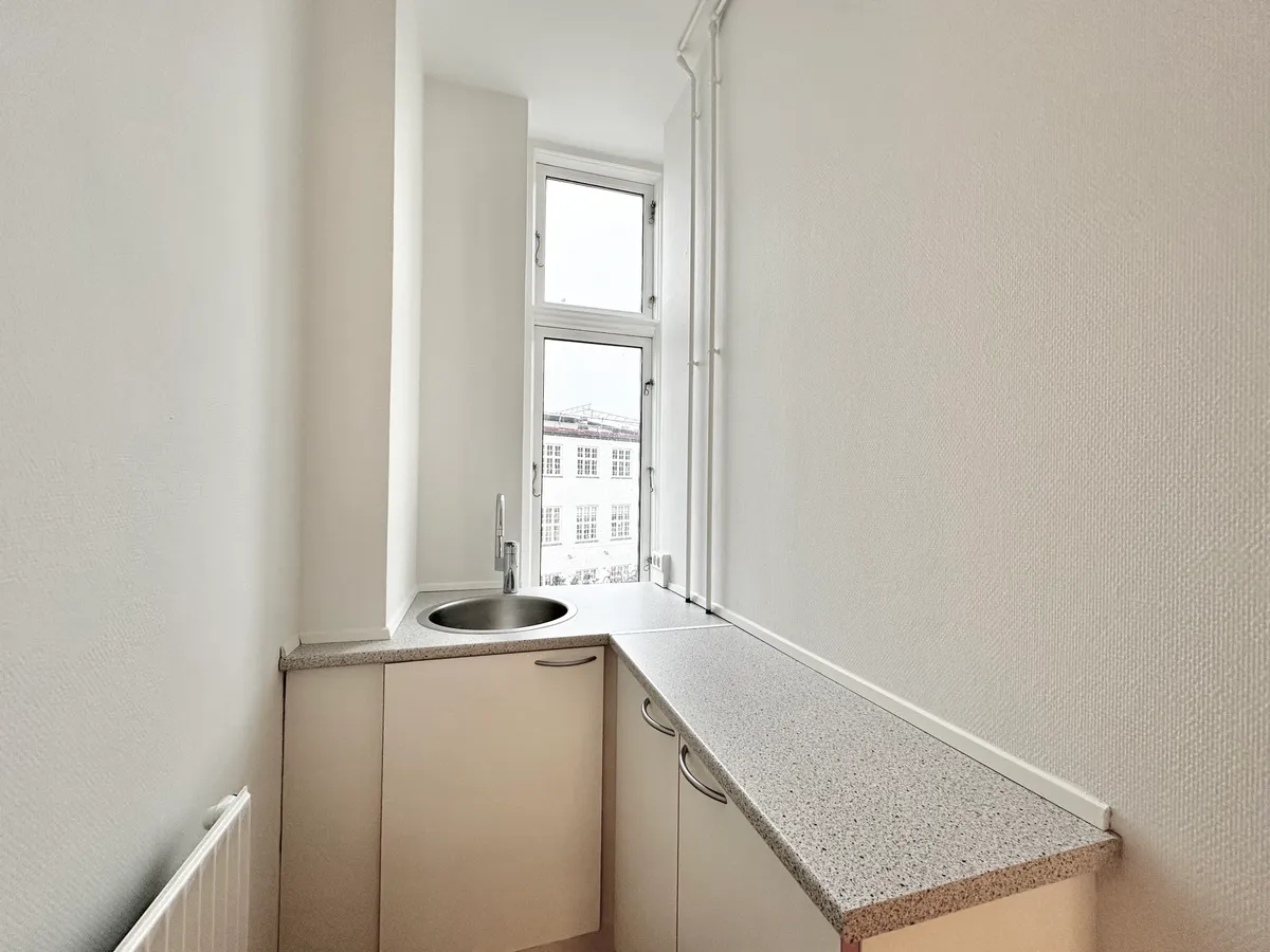 En lille hvid køkkenvæg med en sink, en håndkleholder og et vindue med udsigt til en bygning