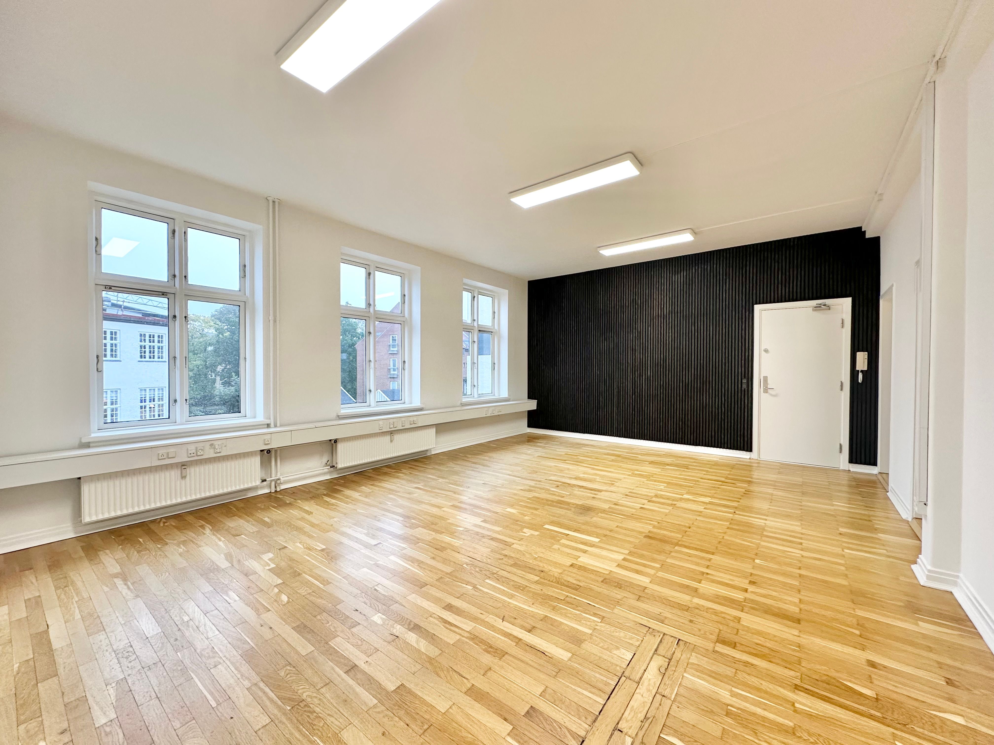 Et moderne kontor med hårdt trægulv, lysevinduer og radiatorer. Et rum med en hvid dør og en sort væg. To lyse paneler er monteret på loftet.