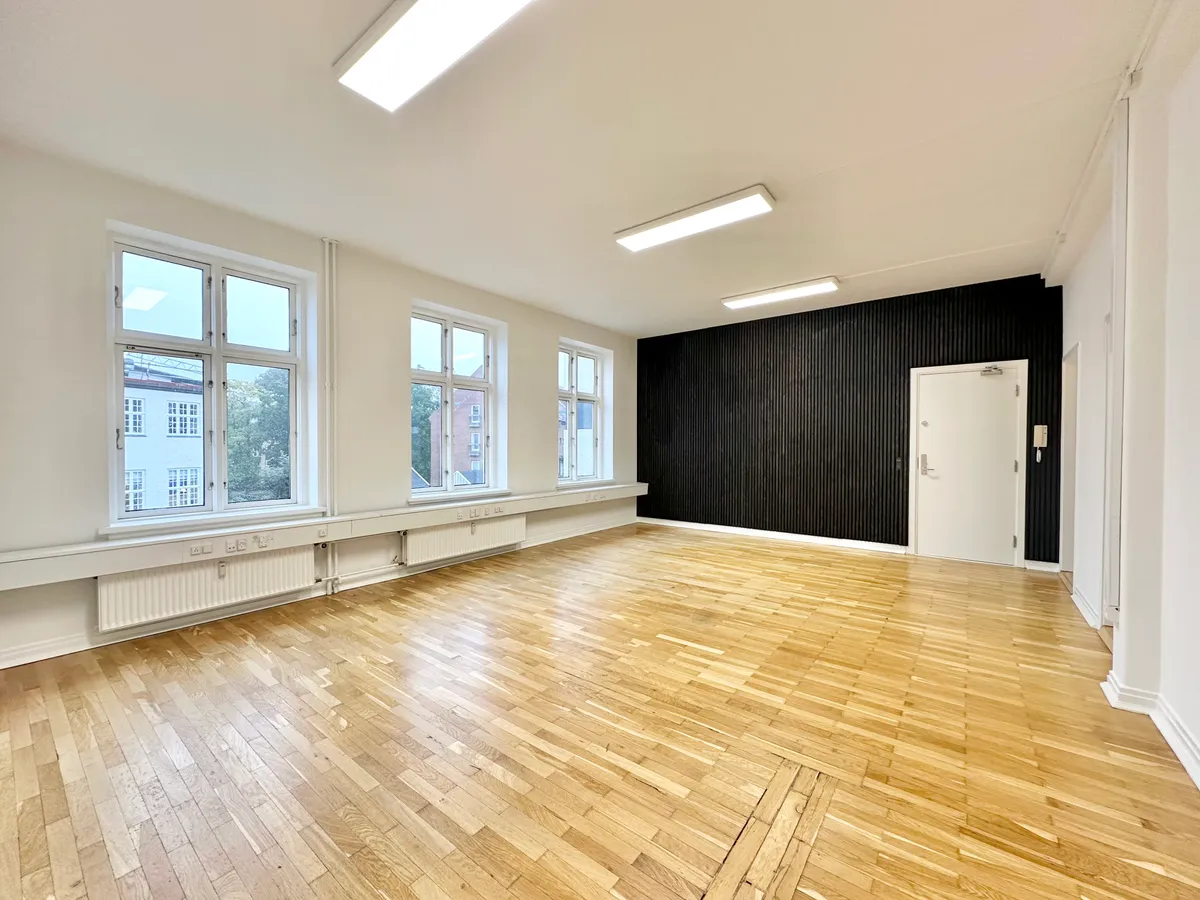 Et moderne kontor med hårdt trægulv, lysevinduer og radiatorer. Et rum med en hvid dør og en sort væg. To lyse paneler er monteret på loftet.