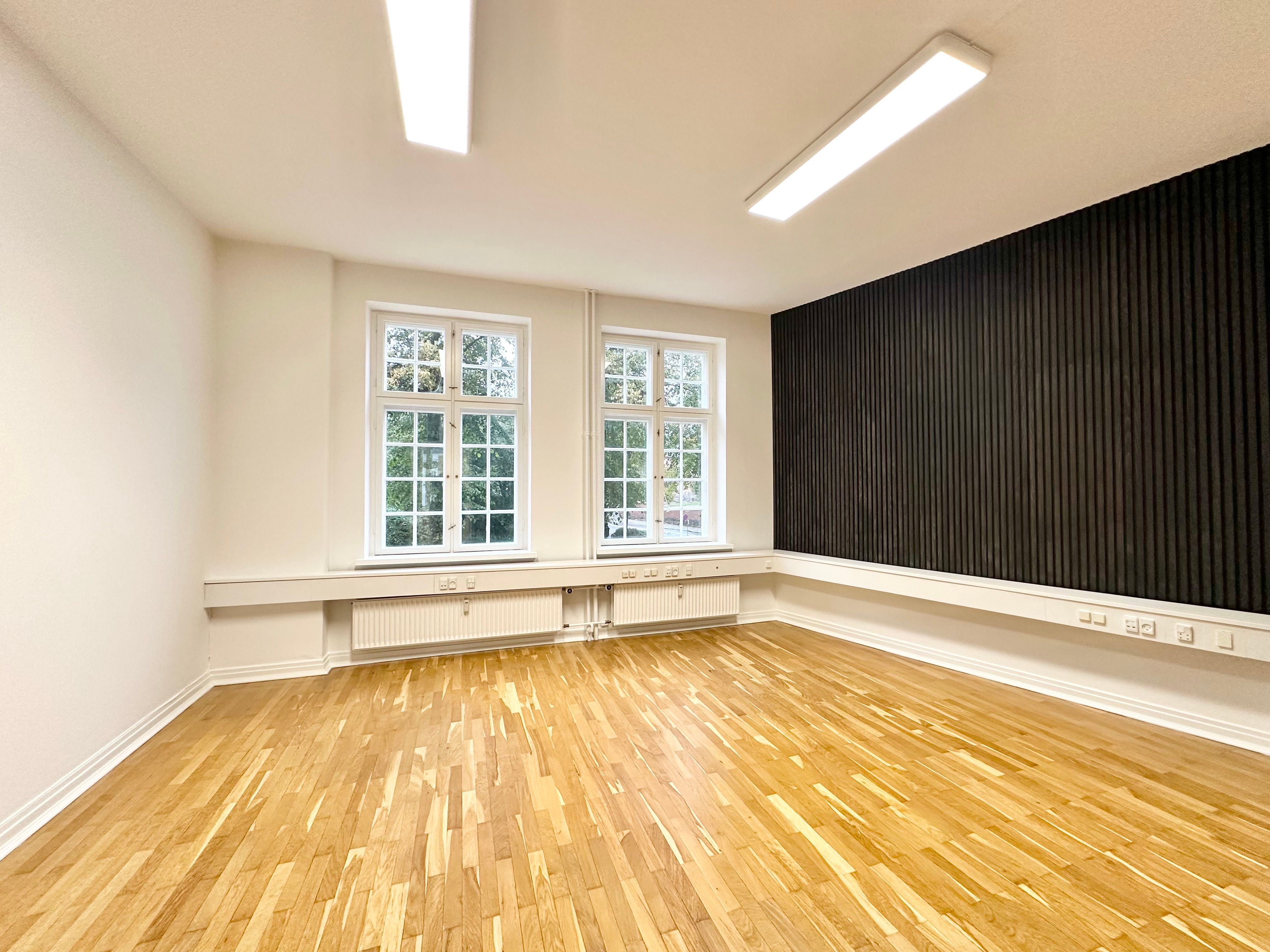 Et moderne og rent kontor med lyse trægulve, to vinduer og hvide vægge. Der er radiatorer under vinduerne og lyse lys på loftet.