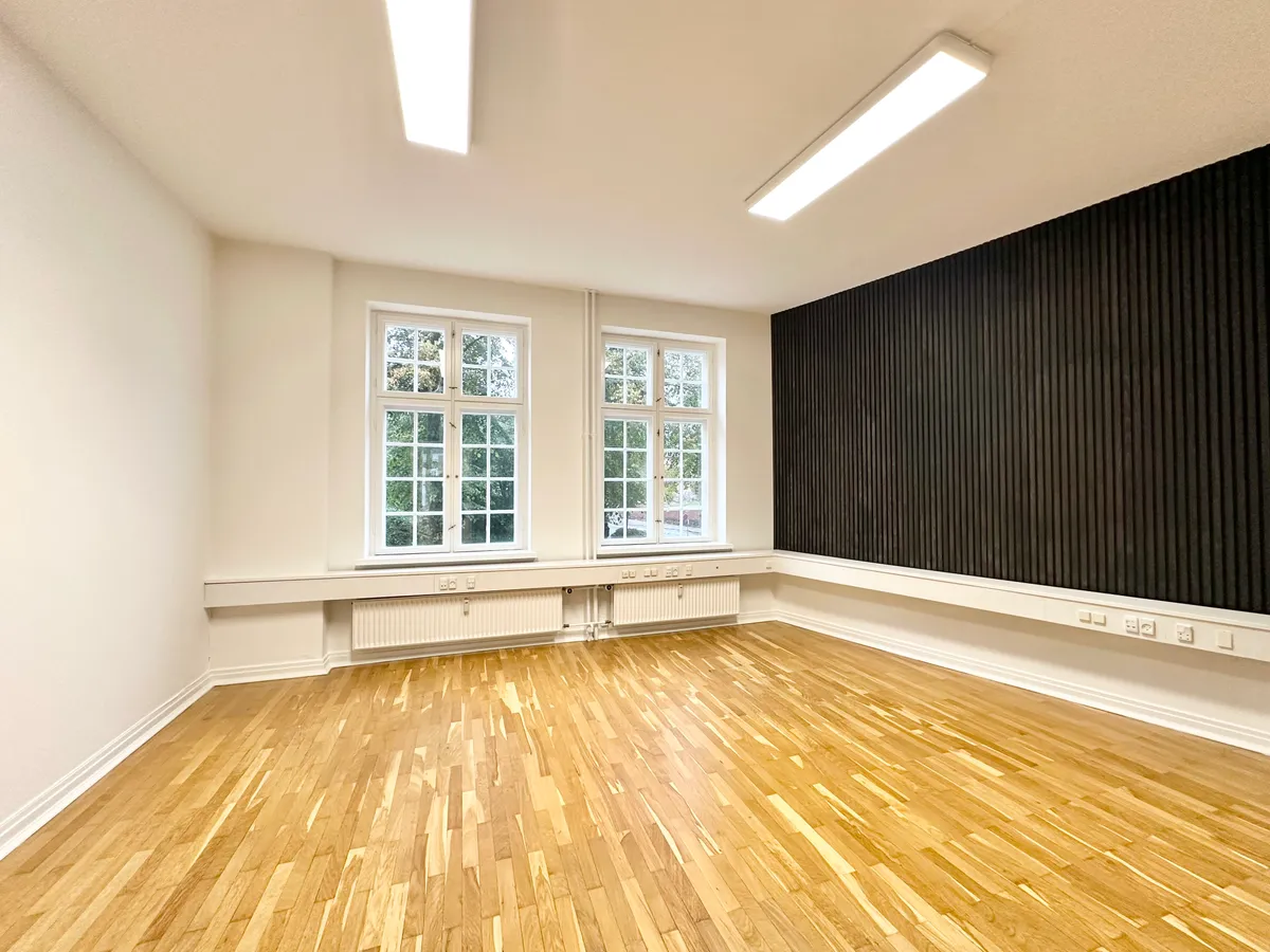 Et moderne og rent kontor med lyse trægulve, to vinduer og hvide vægge. Der er radiatorer under vinduerne og lyse lys på loftet.