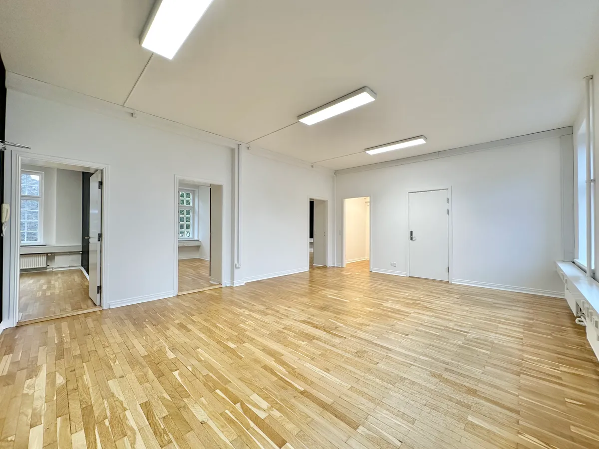 Stort og lyst kontorloft med lyse paneler og tre lysekroner. Lejligheden har lyse parketgulve og hvide vægge. Der er åbne døre og vinduer med udsigt til gaden