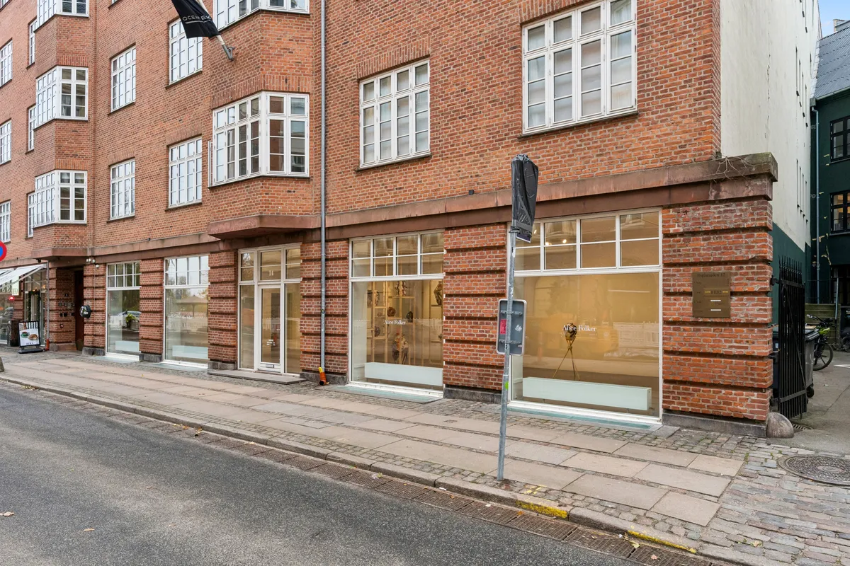 Lejlighed til leje på Esplanaden 14 med glasruder, røde mursten og en skraldspand ved siden af. Butiksvinduer med udstillinger og en væg med plakater