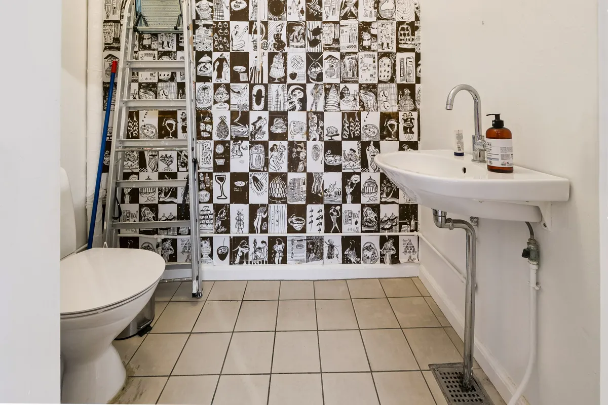 En lille badeværelse med en toilet, en hvid vaskeske og et sæbeflaske på vaskebassinet og et stige med en blå støvsugerbørste i hjørnet