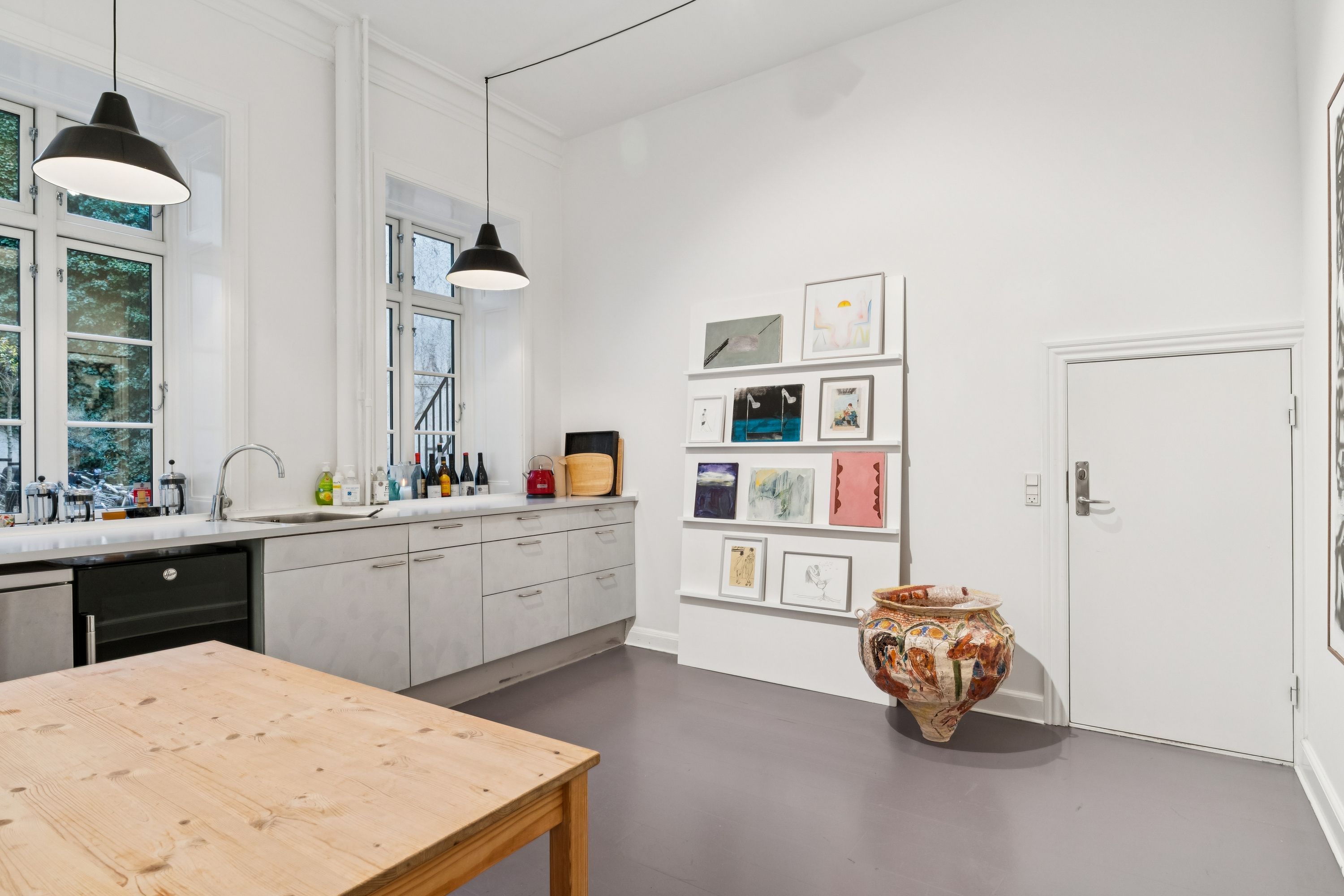 Lyst køkken med moderne møbler og lyse loftslampe. Sort køleskab, vask og lyse loftslamper. Væggen er hvid og der er et værktøjsreol med diverse kunstværker.