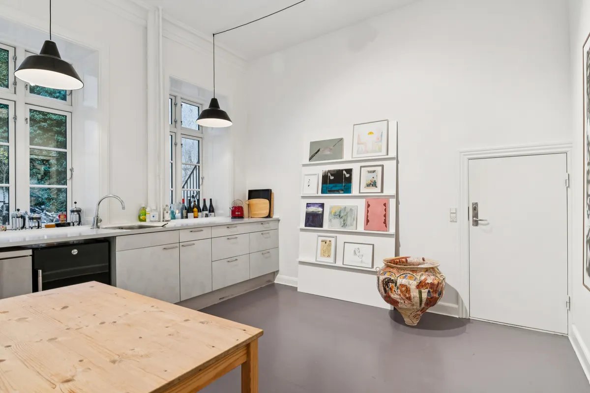 Lyst køkken med moderne møbler og lyse loftslampe. Sort køleskab, vask og lyse loftslamper. Væggen er hvid og der er et værktøjsreol med diverse kunstværker.