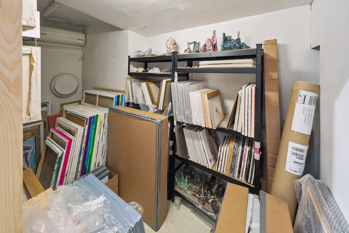 Sort, fyldt opbevaringsrum med høje hylder og masser af papkasser og ruller i gulvet. Store kunstværker, skulpturer og emballagematerialer er spredt rundt.