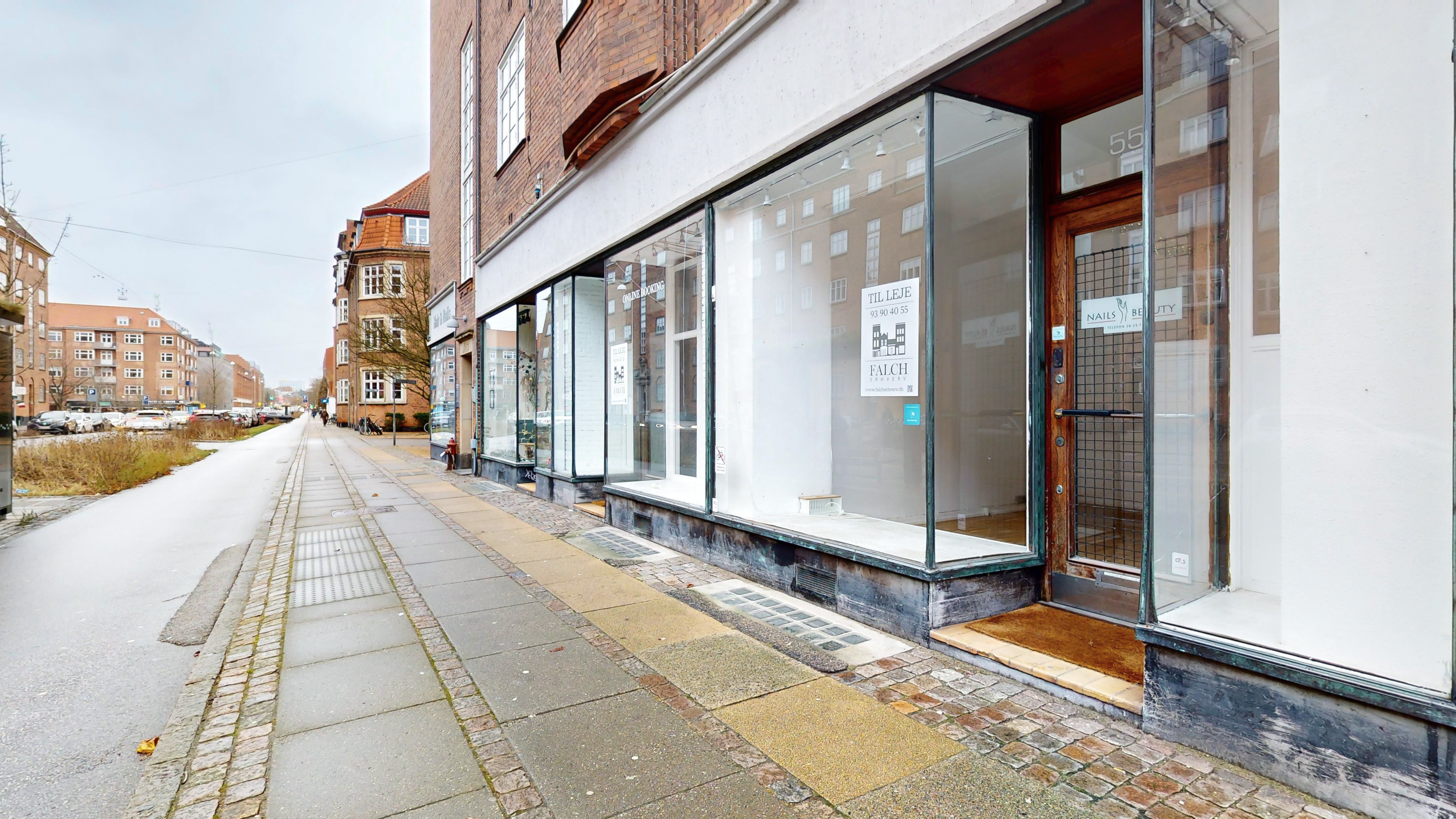 Forretningslokaler til leje på Peter Bangs Vej 53, Frederiksberg. To butikker med glasvinduer og træindgange. Oplysninger om lejen og kontakter står på skilte i vinduer. Parkerede biler og grønne buske langs gaden