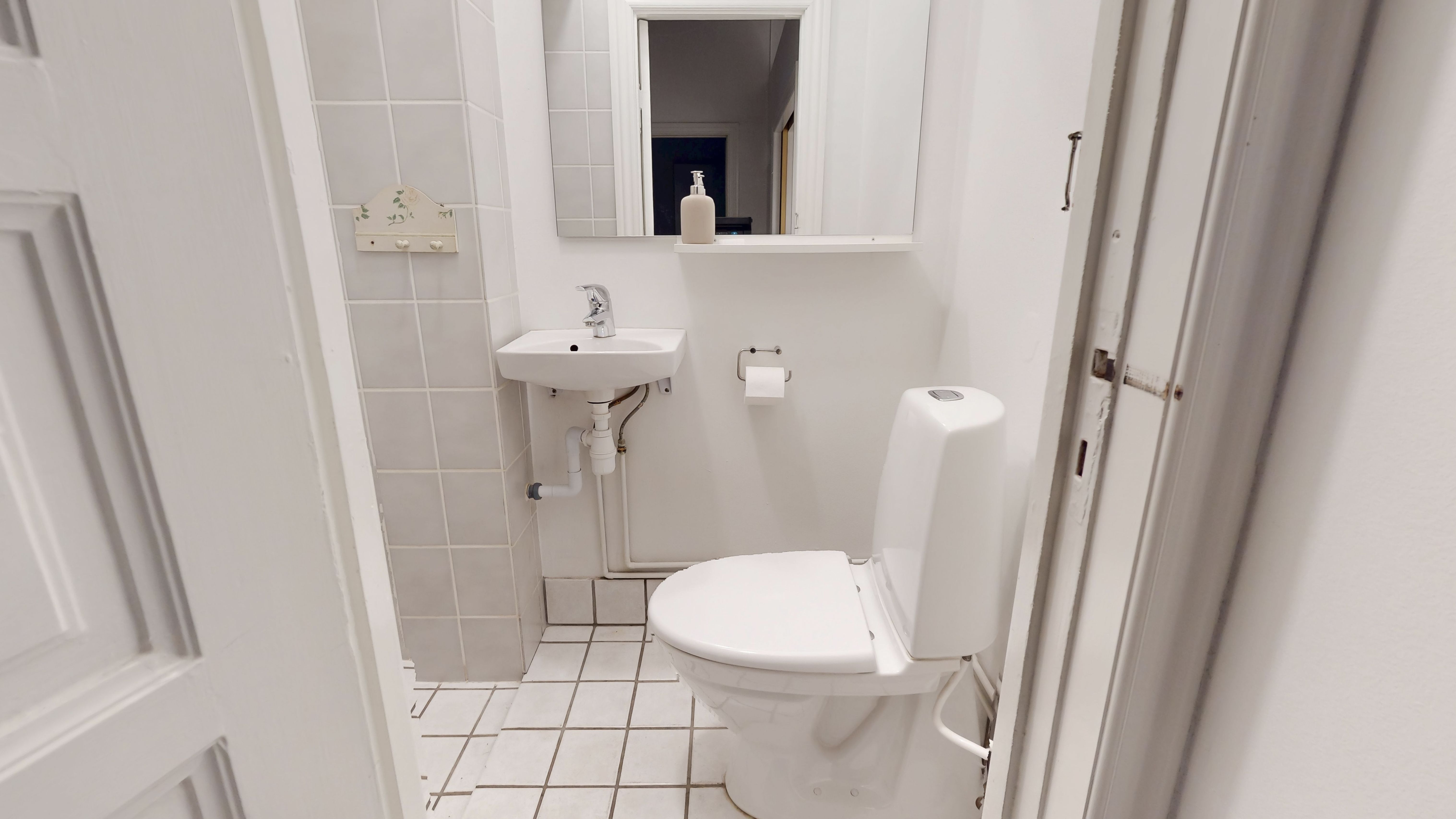 En hvid toilet i et lille badeværelse med en hvid håndvask og et spejl på væggen