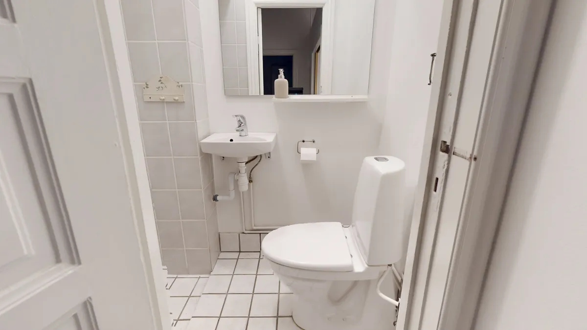 En hvid toilet i et lille badeværelse med en hvid håndvask og et spejl på væggen