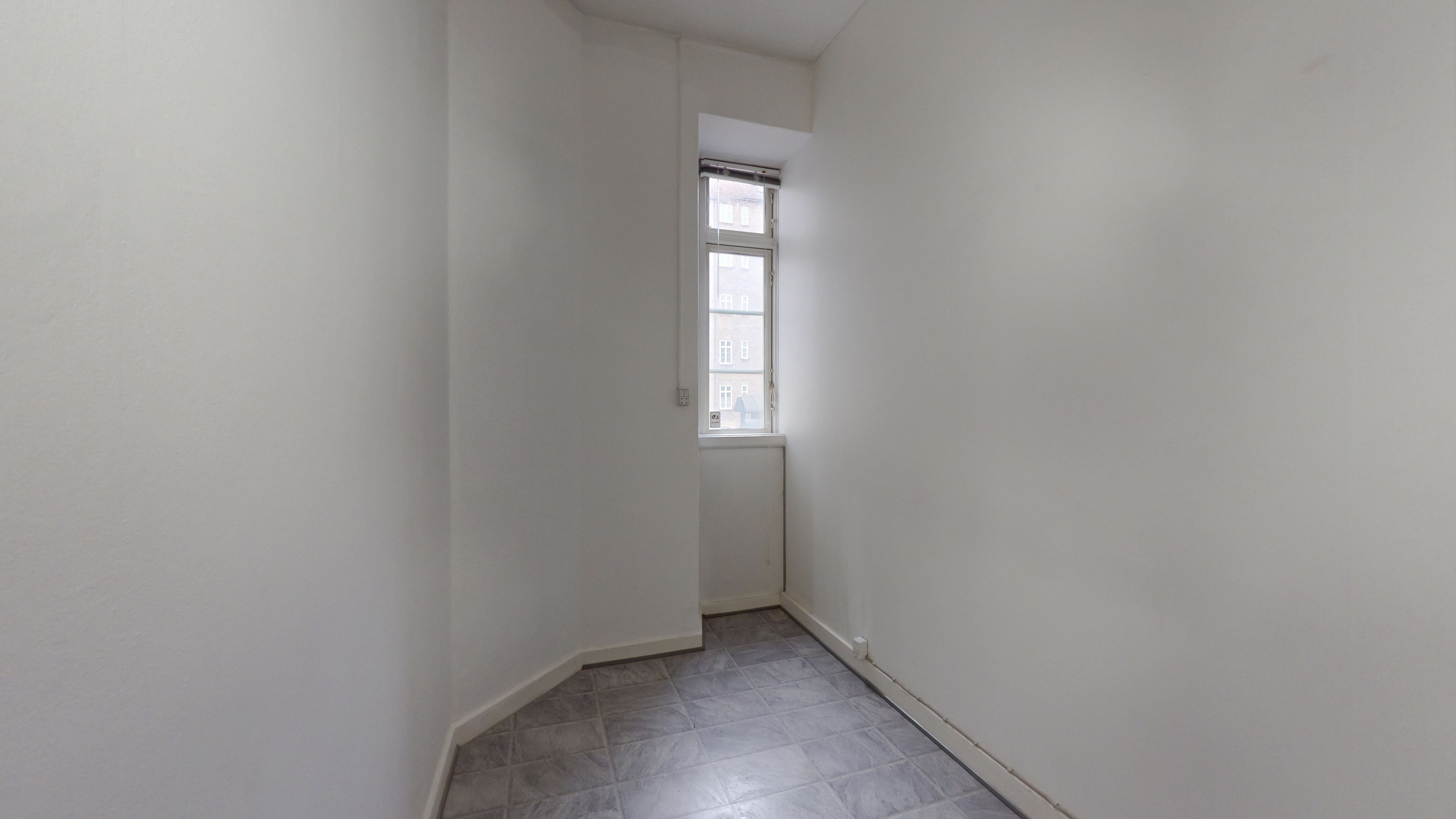 En hvid malet væg med et vindue, der giver udsigt til bygninger og en klar himmel. Gulvet er lavet af grå tømmerklinker.