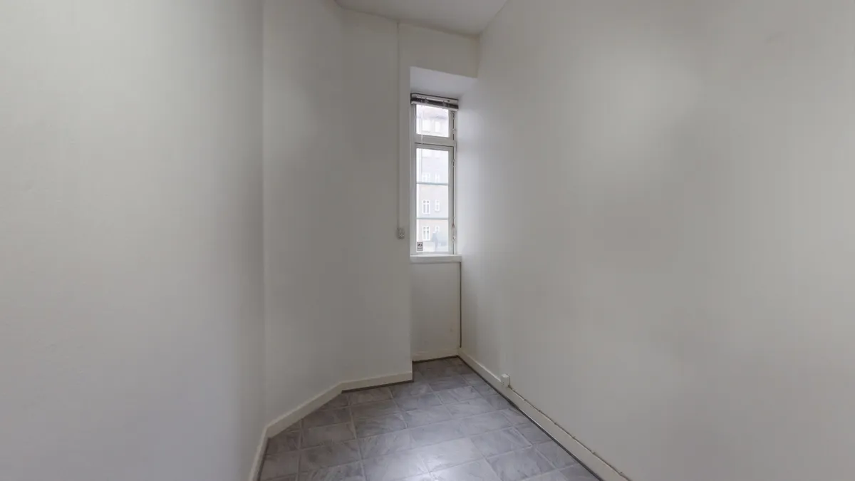 En hvid malet væg med et vindue, der giver udsigt til bygninger og en klar himmel. Gulvet er lavet af grå tømmerklinker.