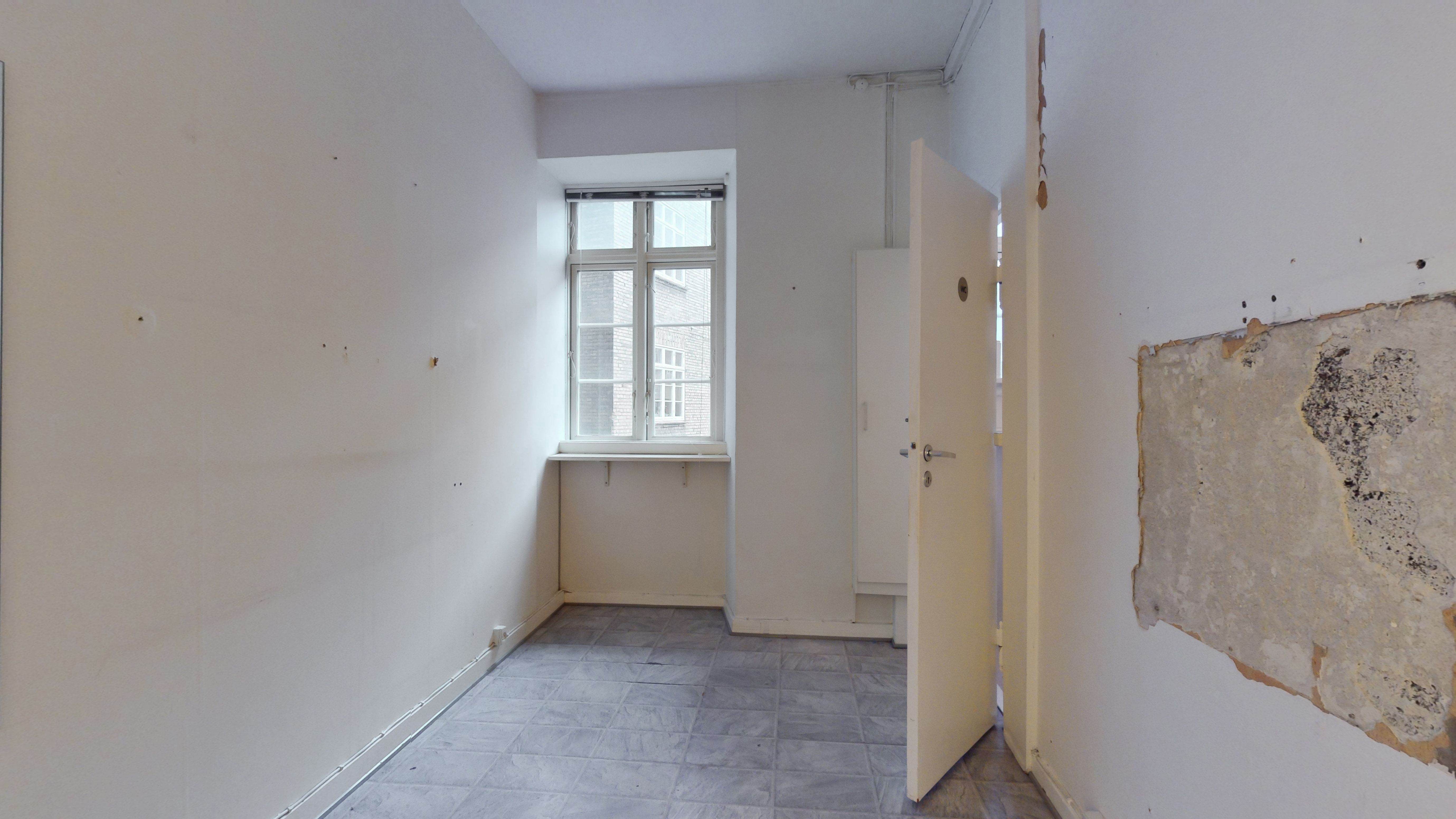 En tom lejlighed med hvide vægge og en åben dør til et andet værelse med en vindueskarm og beskadiget væg.