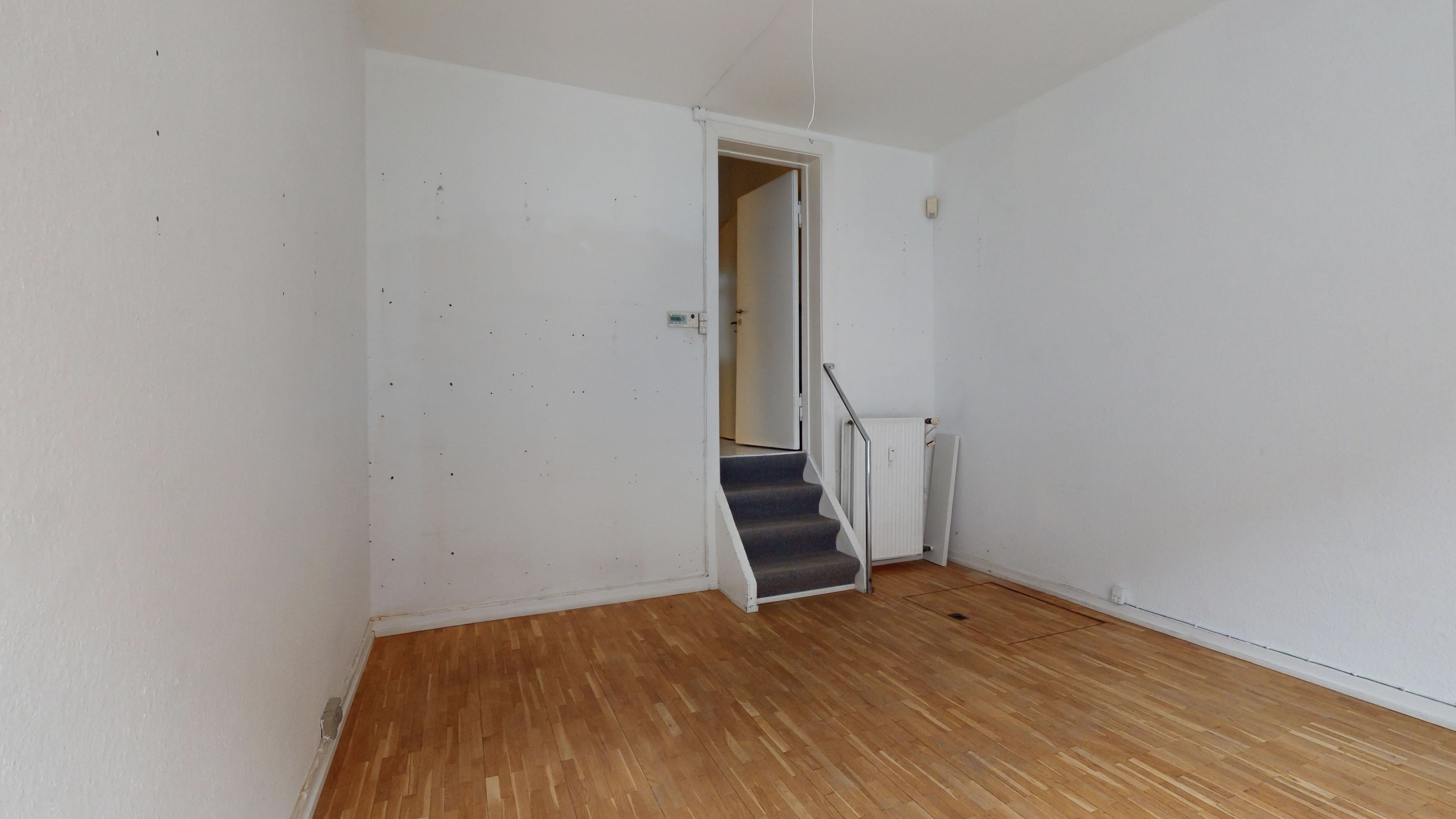 Et tomt værelse med hvide vægge og trægulv, stige og dør i baggrunden, og et radiatorsystem i siden