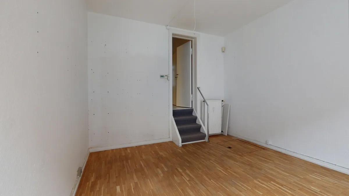 Et tomt værelse med hvide vægge og trægulv, stige og dør i baggrunden, og et radiatorsystem i siden
